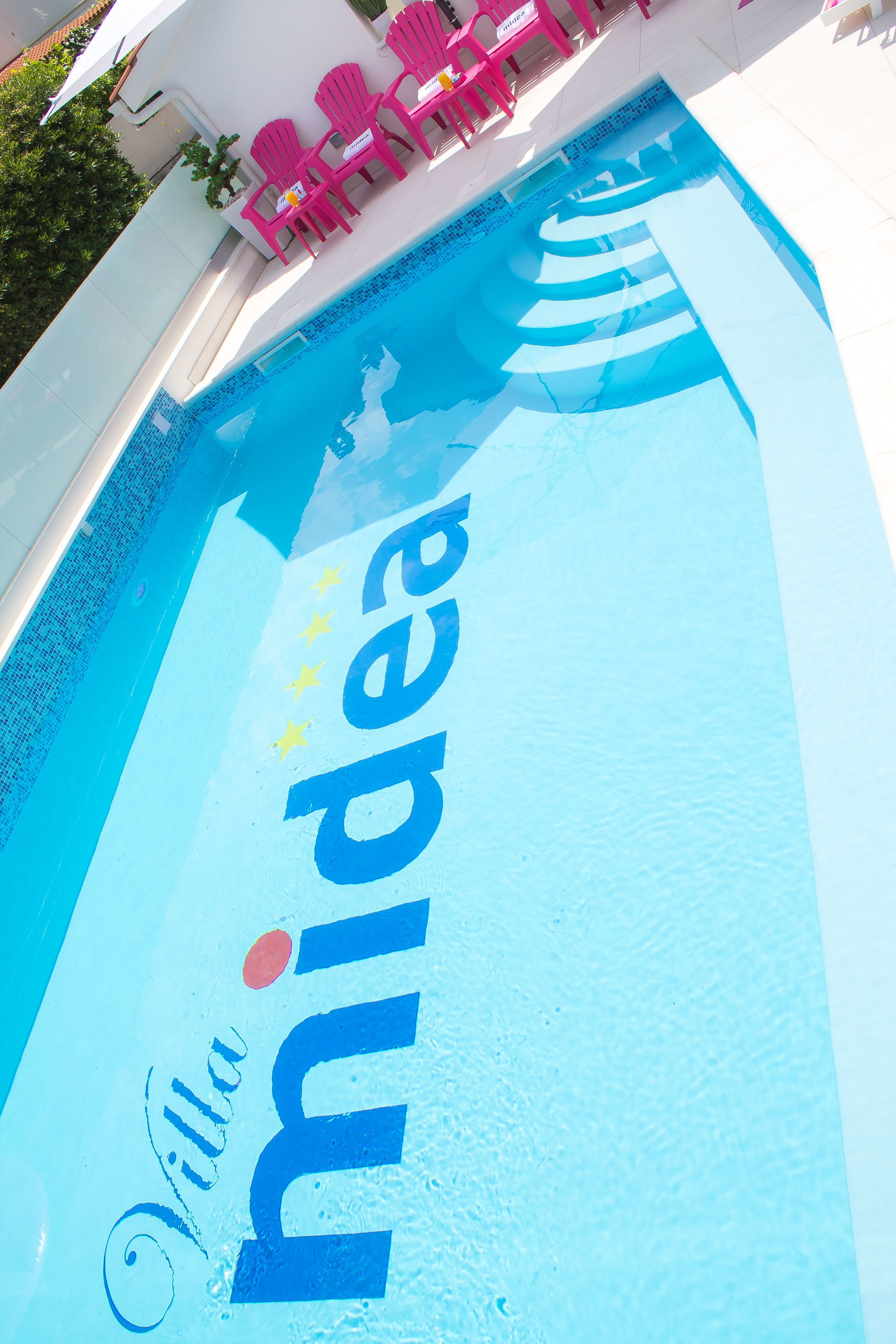 A swimming pool with the text "Villa Midea" and three pink chairs.