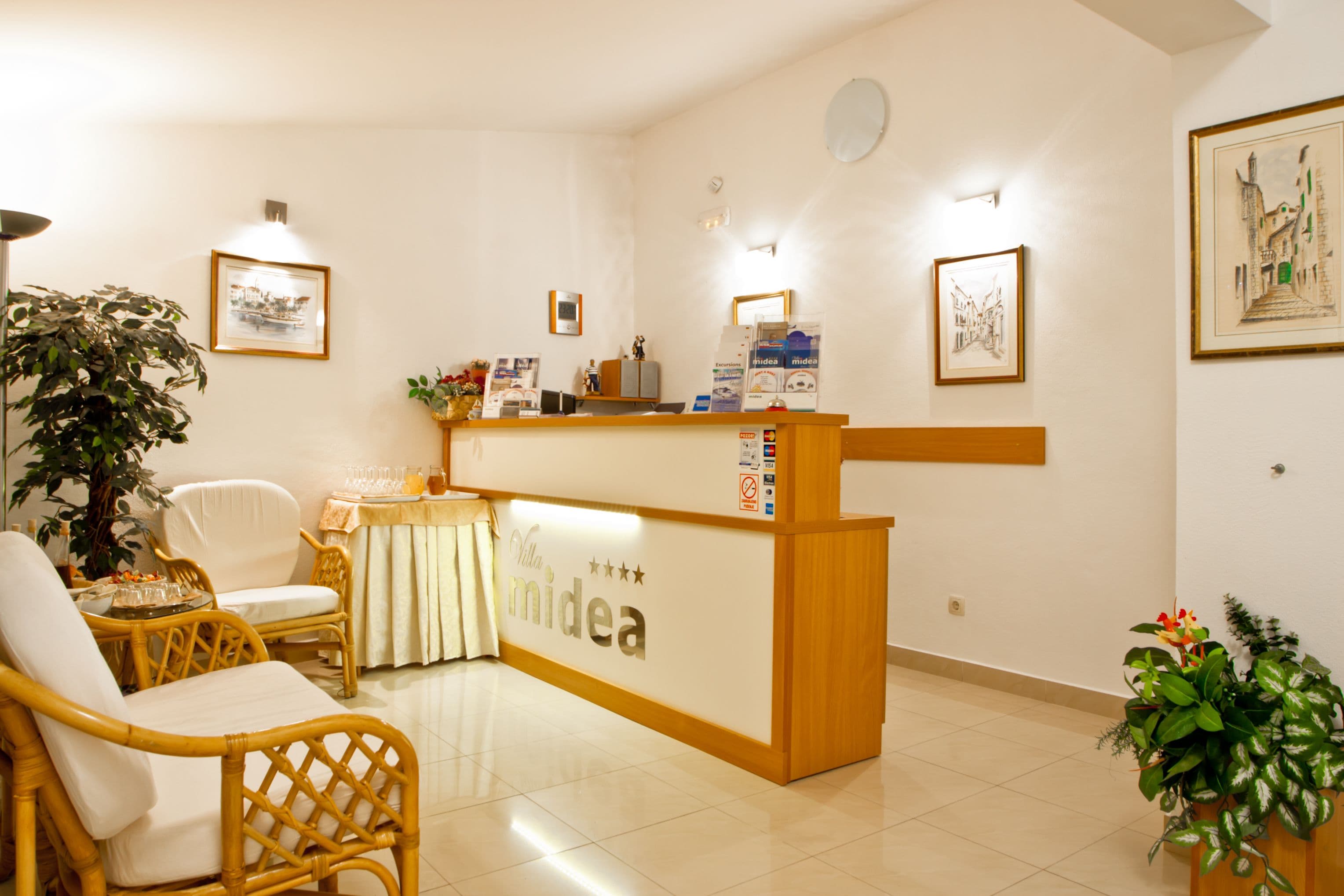 A reception desk with a sign reading "Chillida midea" in a well-lit room with wicker furniture, framed artwork on the walls, and potted plants.