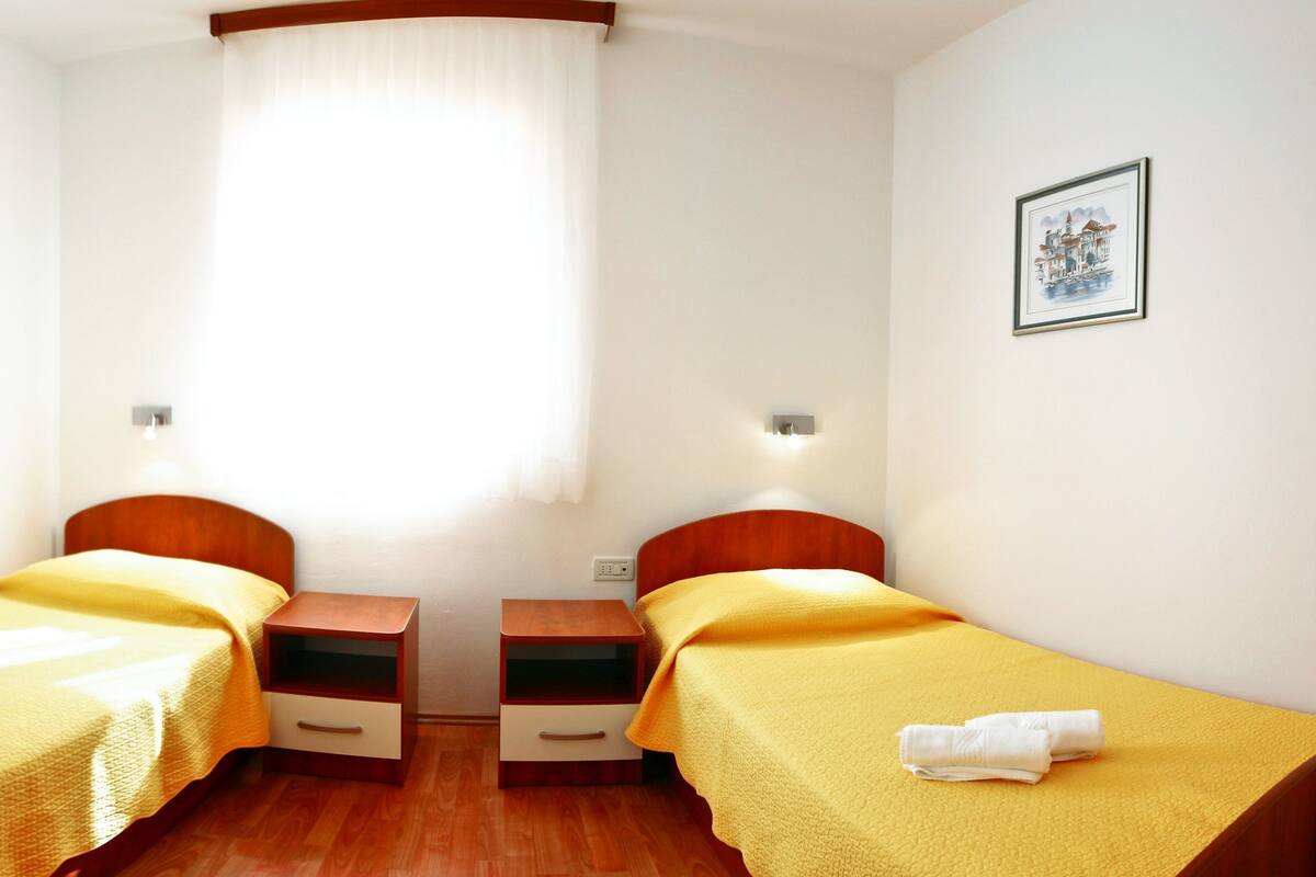 A room with two single beds, wooden headboards, bedside tables with drawers, a window with sheer curtains, and a framed picture on the wall.