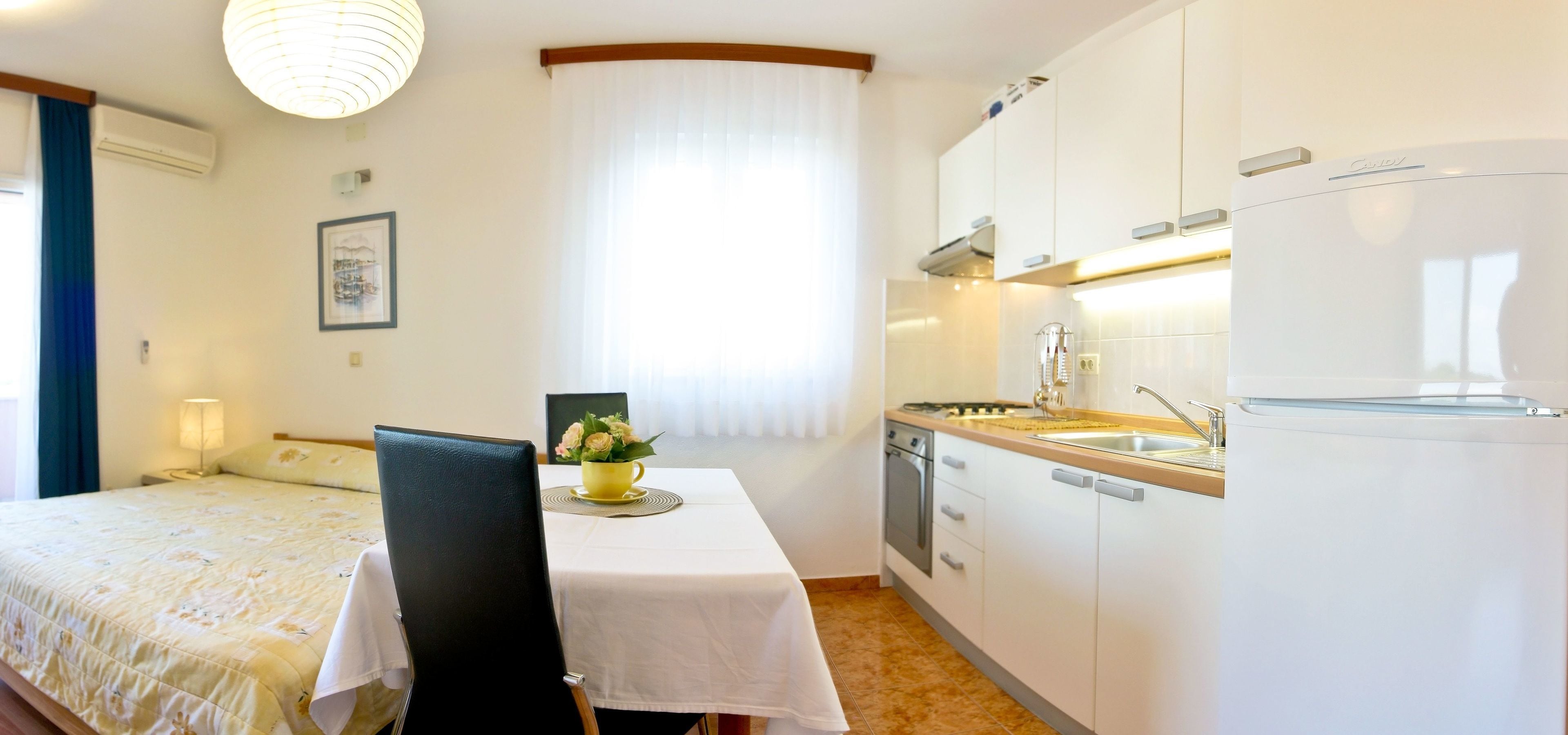 A compact hotel room with a bed, a small dining table with a chair, a kitchen area with a refrigerator, stove, and sink.
