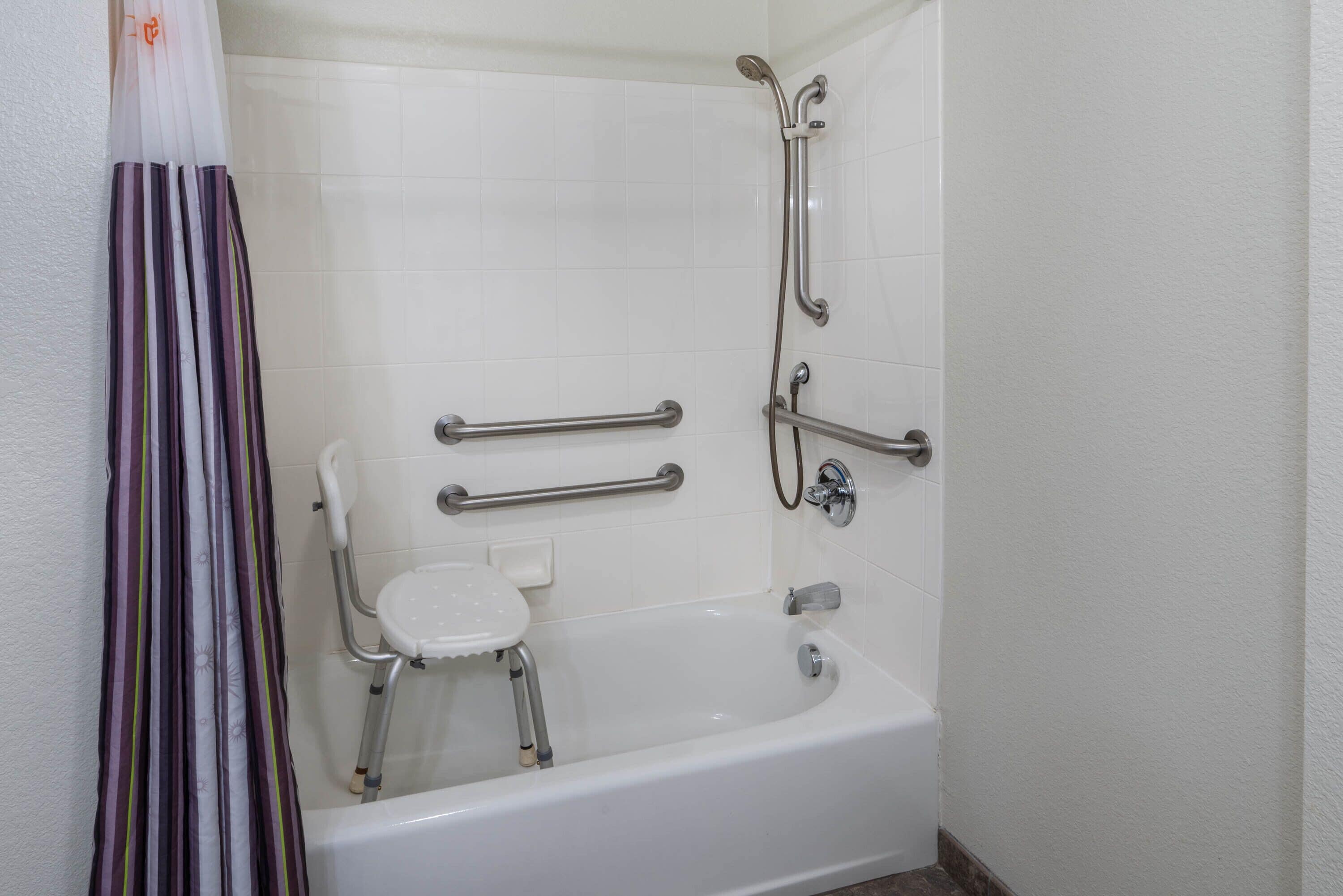 A bathroom with a white bathtub, grab bars, a shower chair, and a handheld showerhead.
