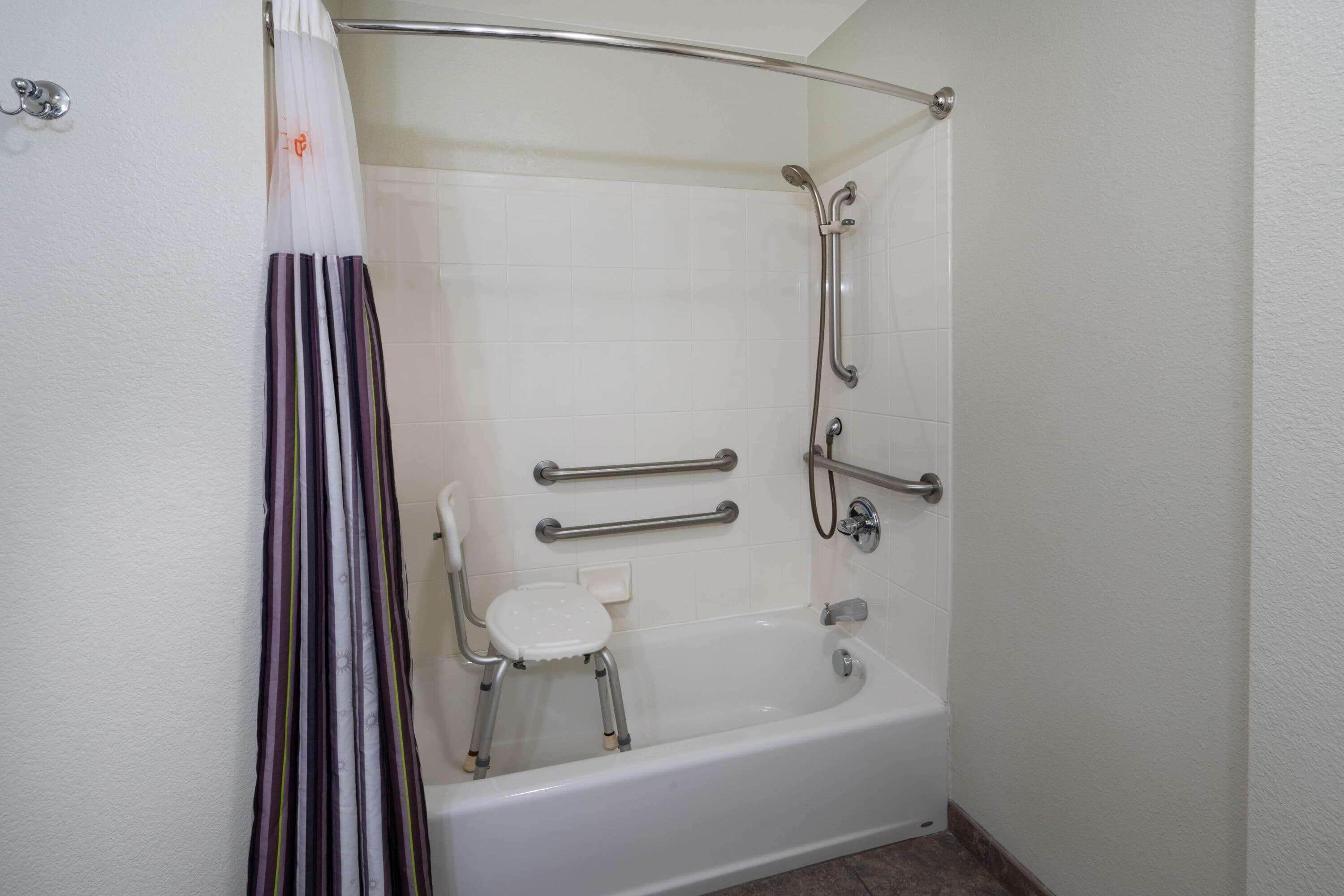 A bathroom with a white bathtub, grab bars, a shower chair, and a shower head.