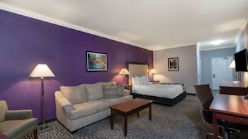 A hotel room with a purple wall, a sofa, a bed, a desk, and two lamps.