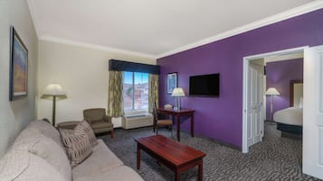 A hotel room with a living area featuring a sofa, a wooden coffee table, a TV, and a window with curtains.