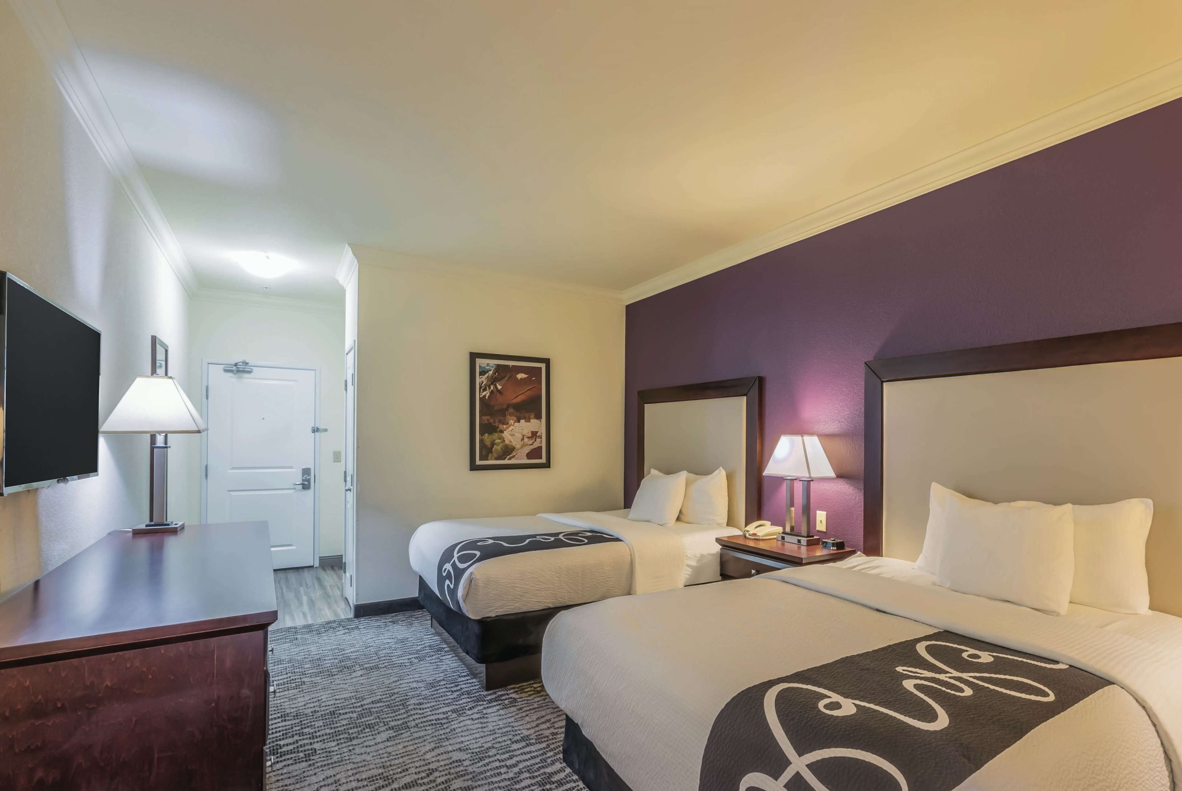 A hotel room with two beds, a desk, a TV, and a door leading to a hallway.