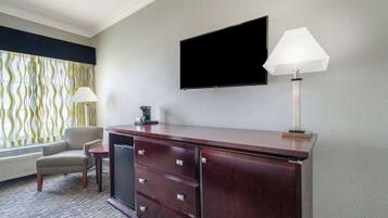 A hotel room with a wooden desk, a flat-screen TV mounted on the wall, a lamp, a chair, and a window with patterned curtains.