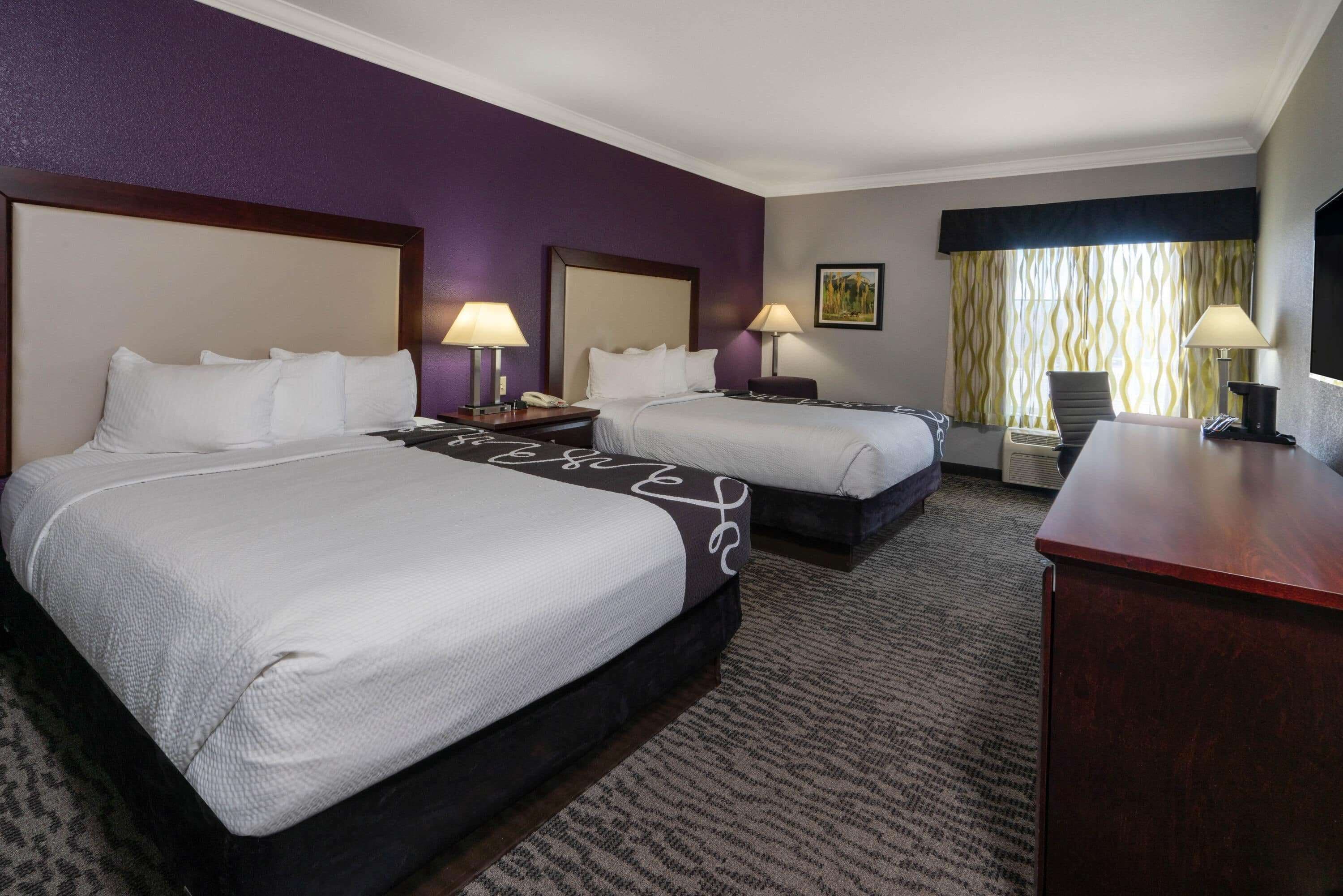 Ein Hotelzimmer mit zwei Betten, einem Schreibtisch und einem Stuhl. Auf dem Schreibtisch stehen Lampen und an beiden Betten Nachttische.