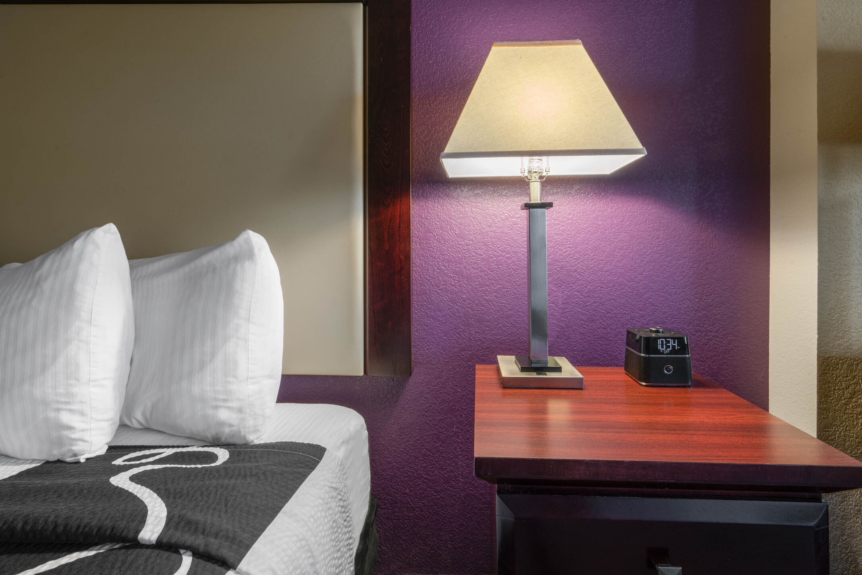 A hotel room with a bed, a nightstand, a lamp, and a digital clock.