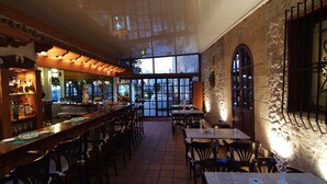A restaurant with a long counter, multiple tables, and chairs. There is a bar area with shelves of bottles and glasses. Large windows provide a view of the outside. - Recó de Randa (Algaida)
