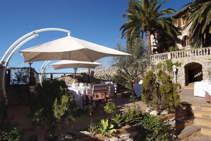 An outdoor dining area with tables and chairs, surrounded by plants and palm trees, under white umbrellas.