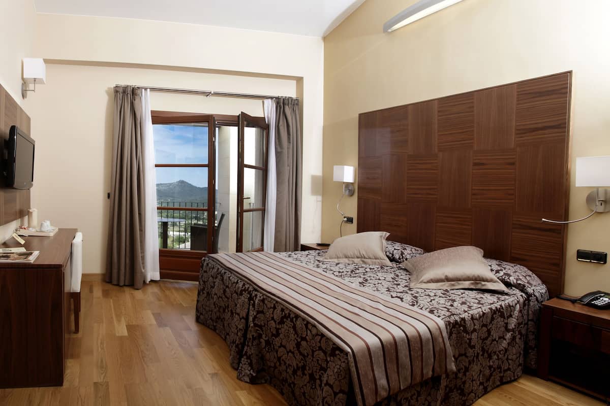 A hotel room with a bed, a desk, a TV, a window with curtains, and a view of the outdoors.