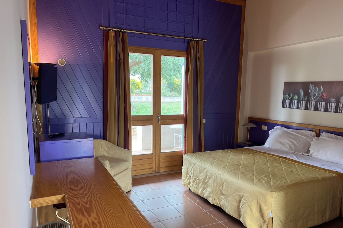 A bedroom with a bed, a desk with a chair, a television, and a window with curtains.