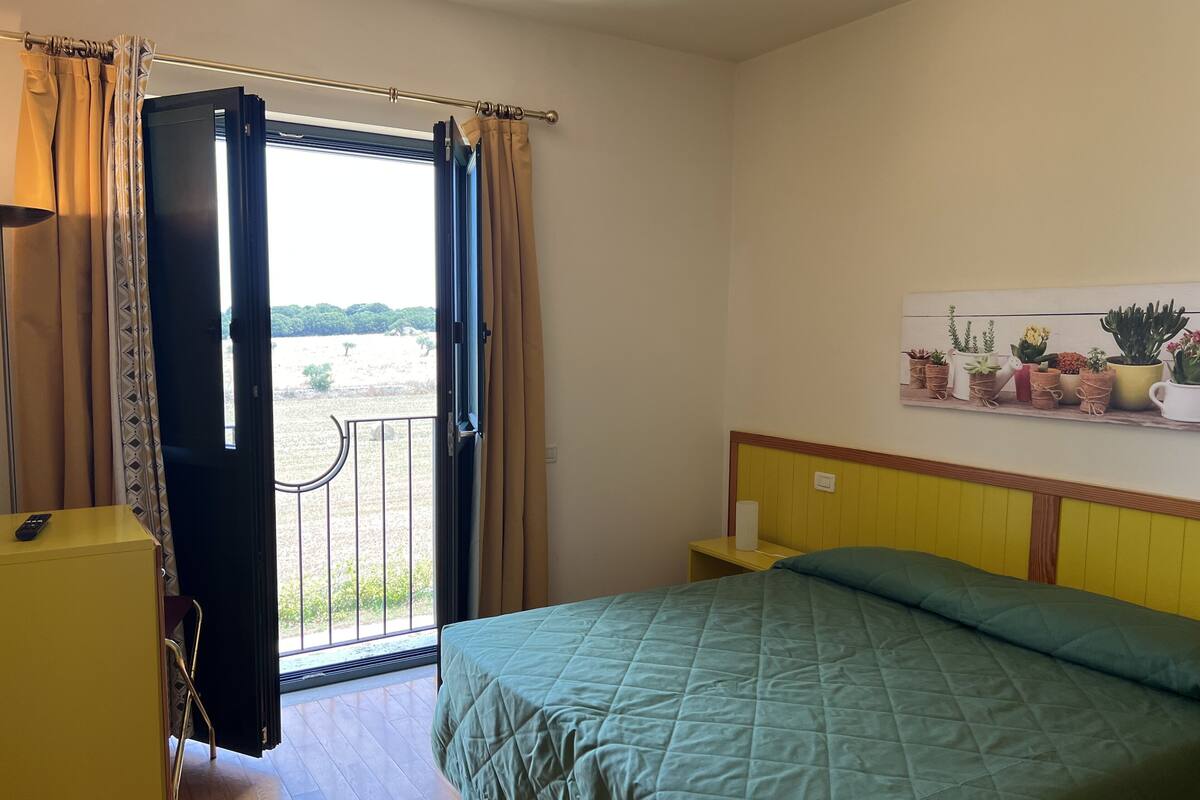 A bedroom with a bed, a nightstand, a painting of plants, and a balcony with curtains.