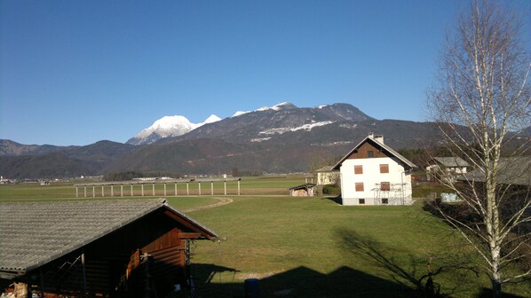 A rural landscape with a white house, a wooden barn, and a mountain with snow-capped peaks in the background.