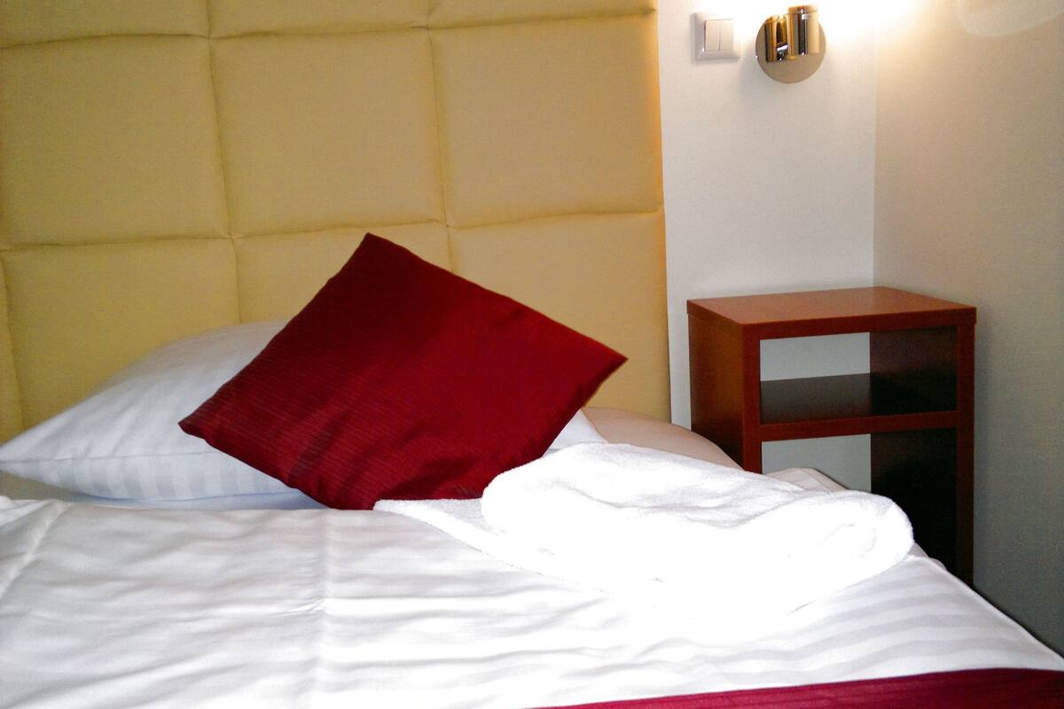 A bed with a red pillow, white sheets, and a yellow headboard. There is a small wooden nightstand beside the bed.