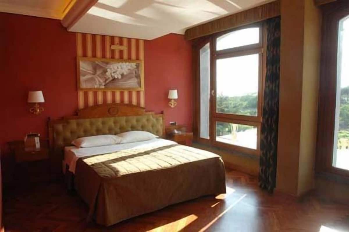 Een slaapkamer met een groot bed, een houten hoofdbord, twee nachtkastjes met lampen en een groot raam met uitzicht op groen.