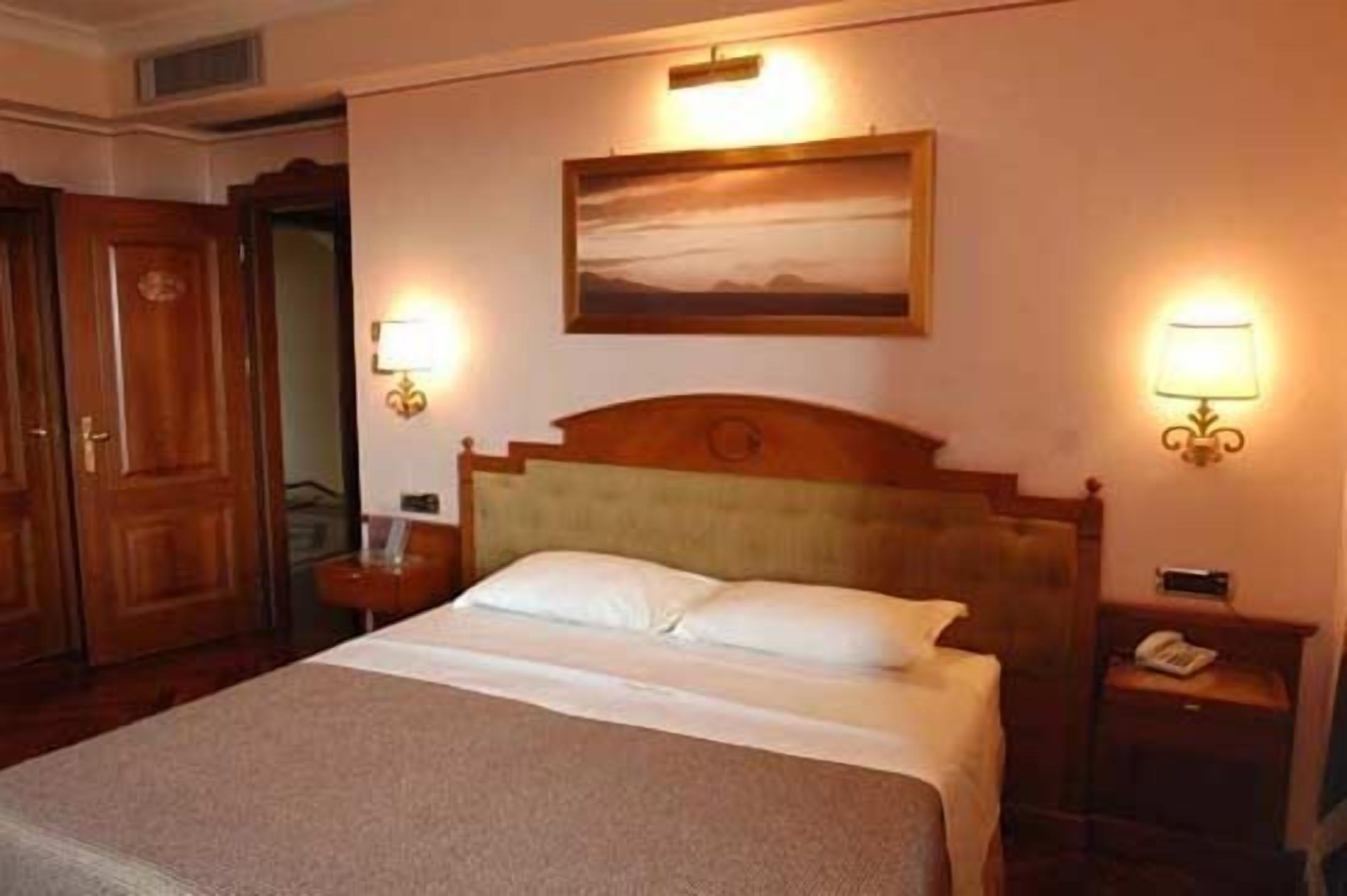 A hotel room with a bed, two bedside tables, a nightstand, a wall-mounted lamp, and a painting above the bed.