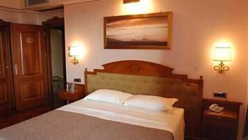 A hotel room with a bed, two bedside tables, a nightstand, a wall-mounted lamp, and a painting above the bed.