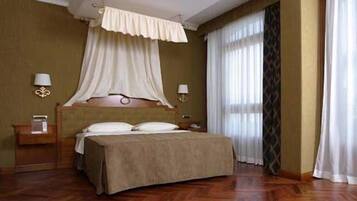 A hotel room with a wooden floor, a bed with a headboard, two bedside tables with lamps, and a window with sheer curtains.