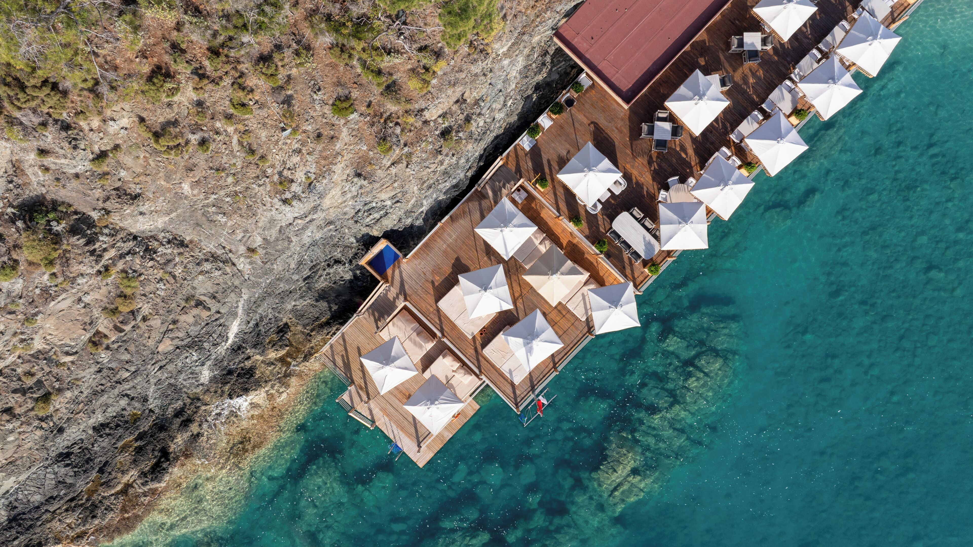 Vue aérienne d’une jetée en bois s’étendant dans des eaux turquoise et limpides, agrémentée de plusieurs parasols blancs et d’espaces aménagés pour s’asseoir.