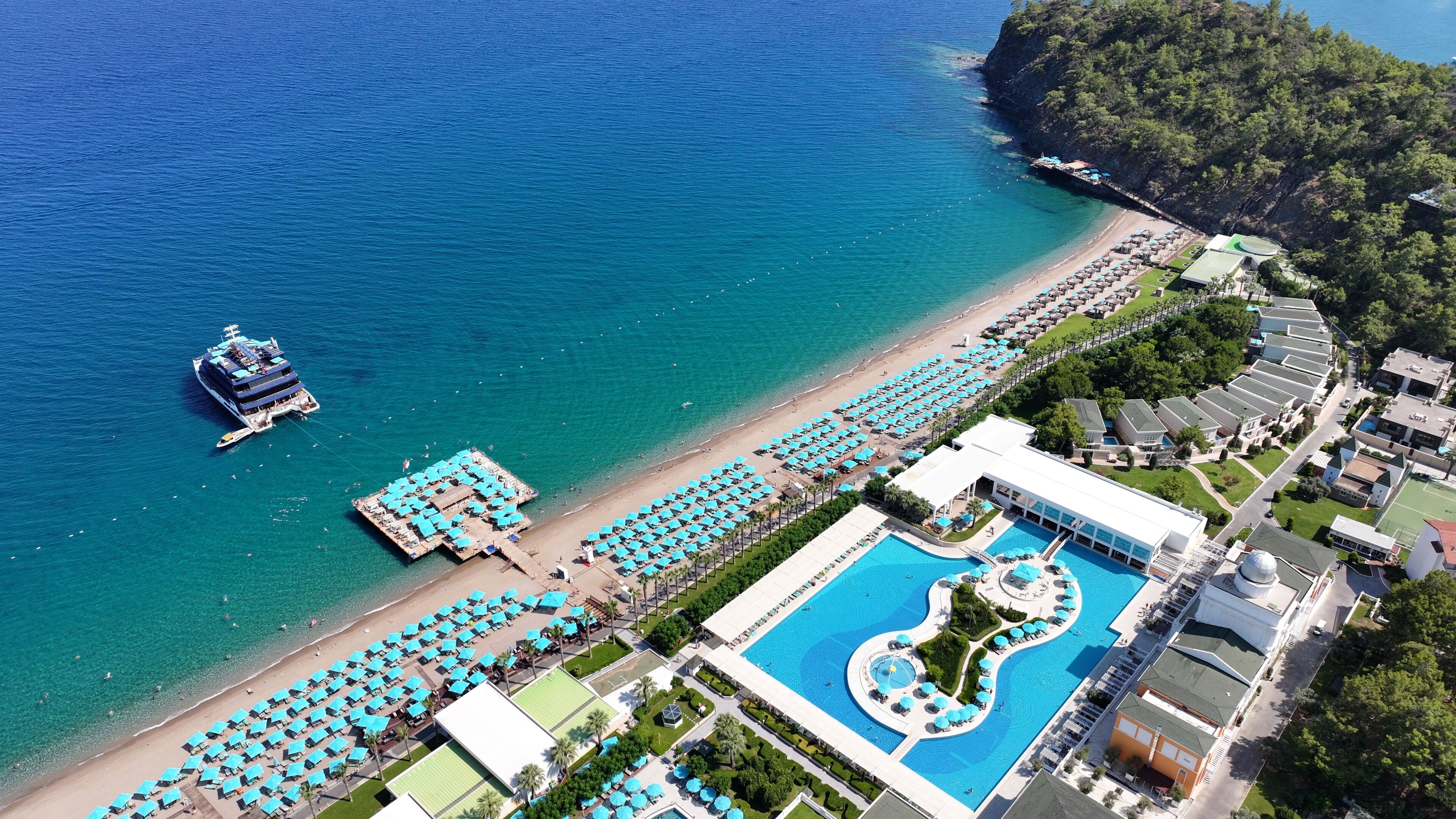 Une vue aérienne d’un complexe hôtelier doté d’une grande piscine, de parasols de plage et d’un yacht amarré près du rivage.