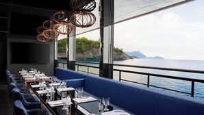 Un restaurante con una larga mesa dispuesta para comer, un banco acolchado azul y vistas al mar.