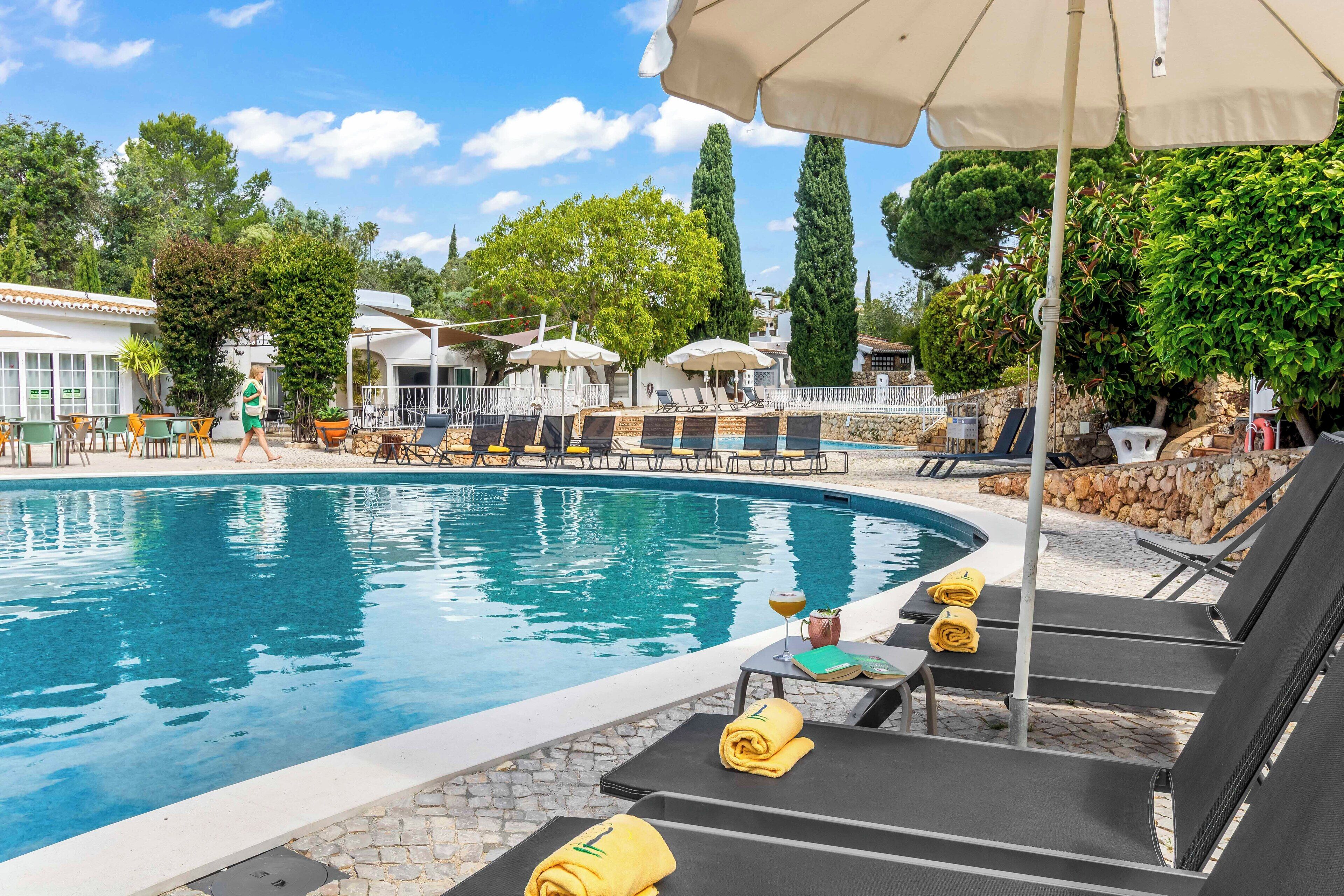 A pool area with lounge chairs and umbrellas, surrounded by trees and a stone wall.