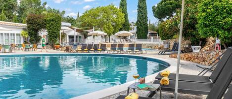 A pool area with lounge chairs and umbrellas, surrounded by trees and a stone wall.