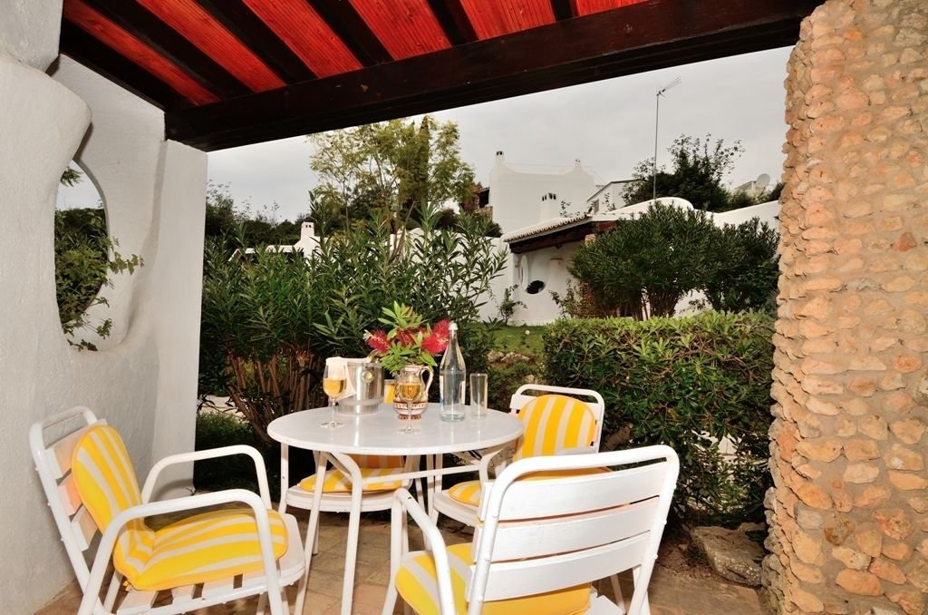 An outdoor patio with a round table and four chairs, surrounded by greenery and a stone wall.