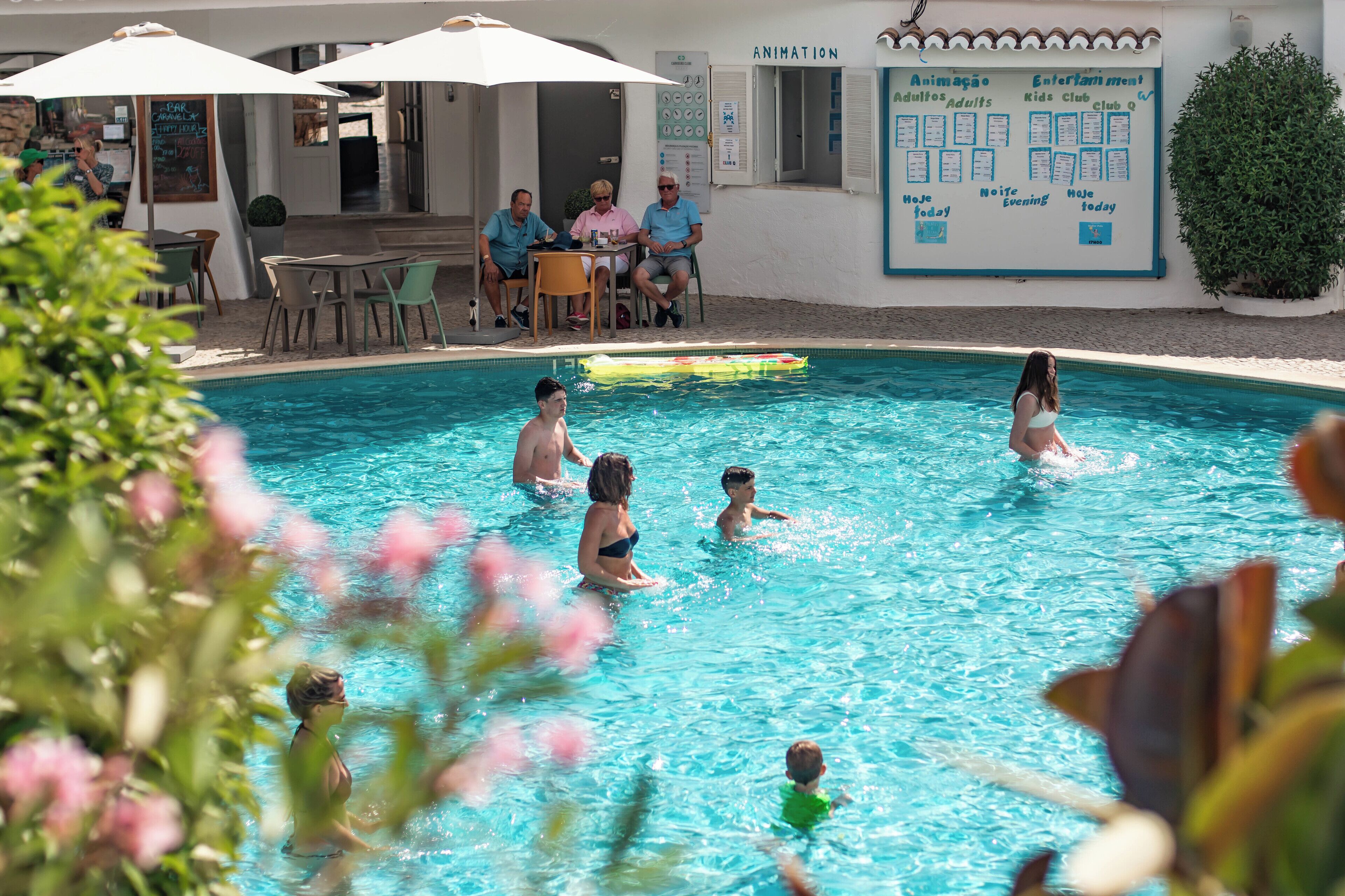 Une piscine avec des personnes dans l’eau, une table avec des chaises et un panneau indiquant différentes options.