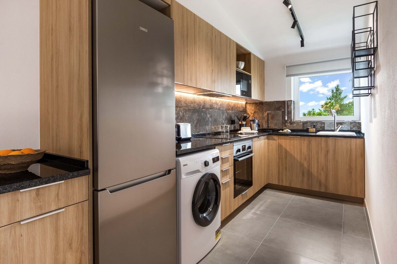 A modern kitchen with wooden cabinets, a stainless steel refrigerator, a washing machine, and a window providing a view of the outdoors.