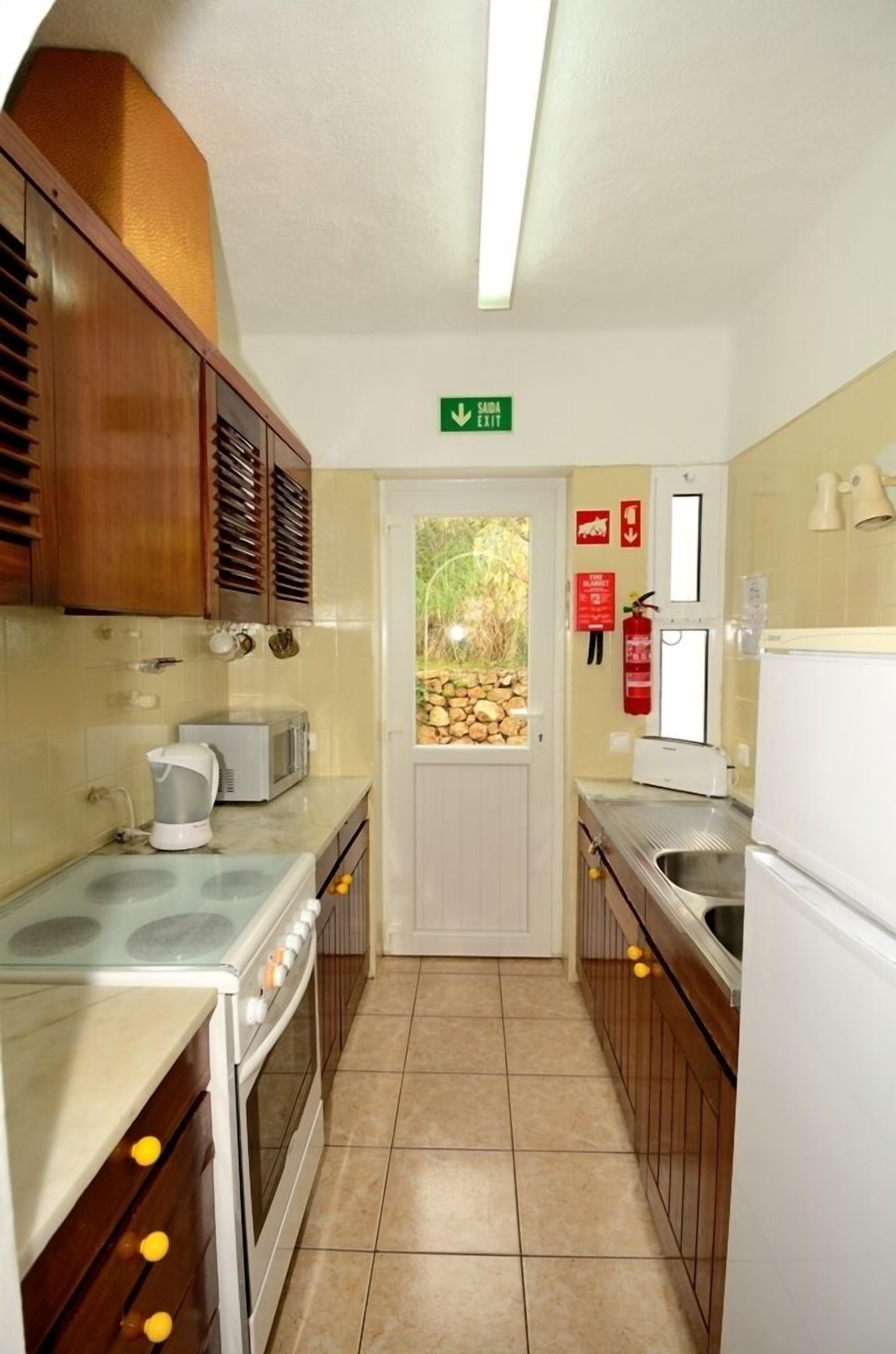 A compact kitchen with a stove, microwave, and sink. There's a door with a green exit sign above it.