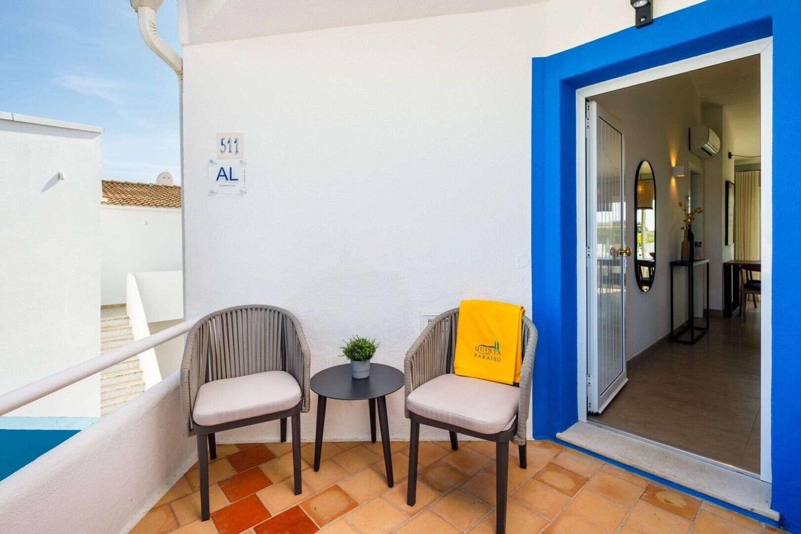 A balcony with two chairs, a small table, and a potted plant. A sign with the number 511 and the letters "AL" is visible on the wall.