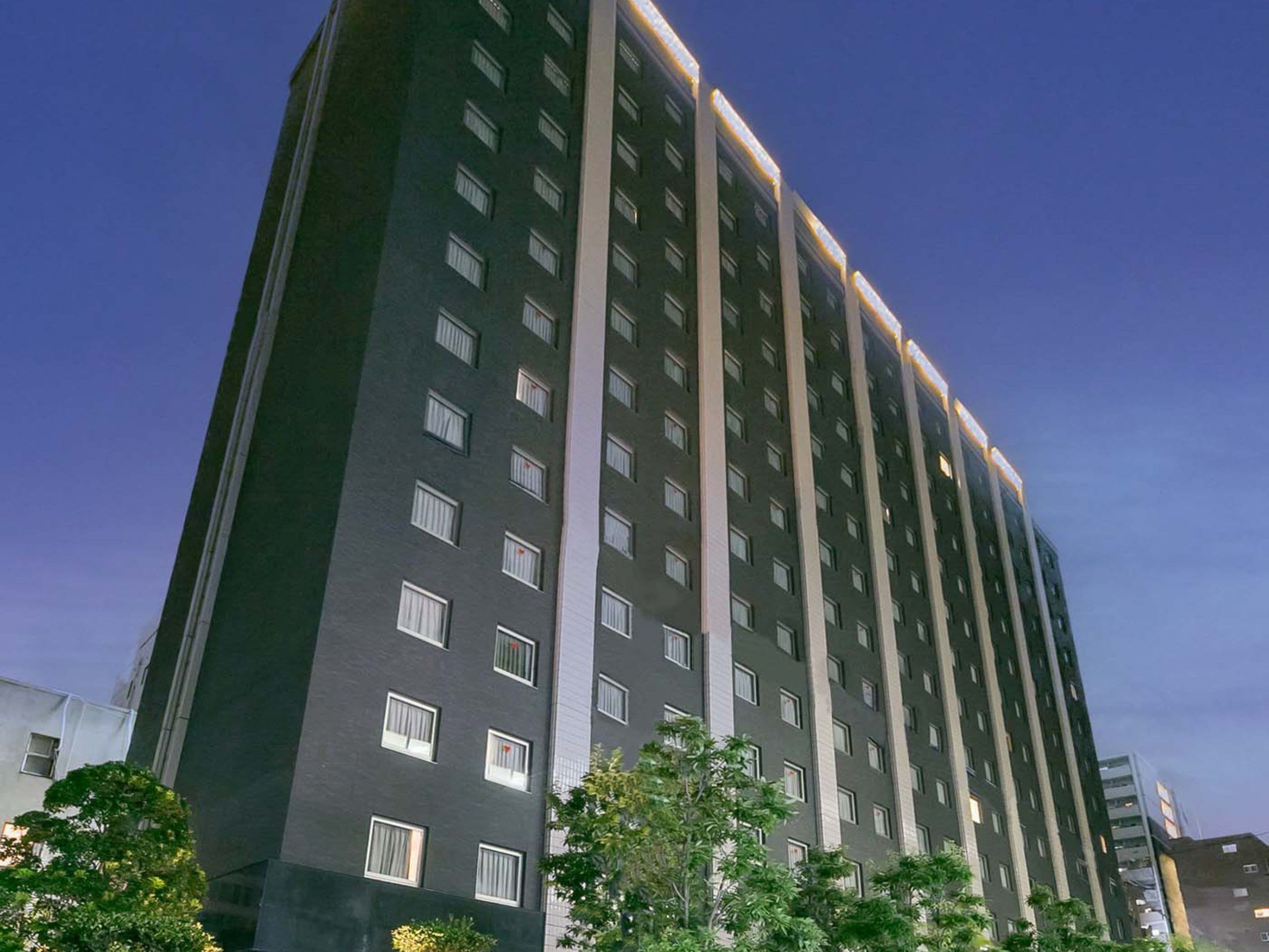 A high-rise building with multiple floors and windows, surrounded by greenery.