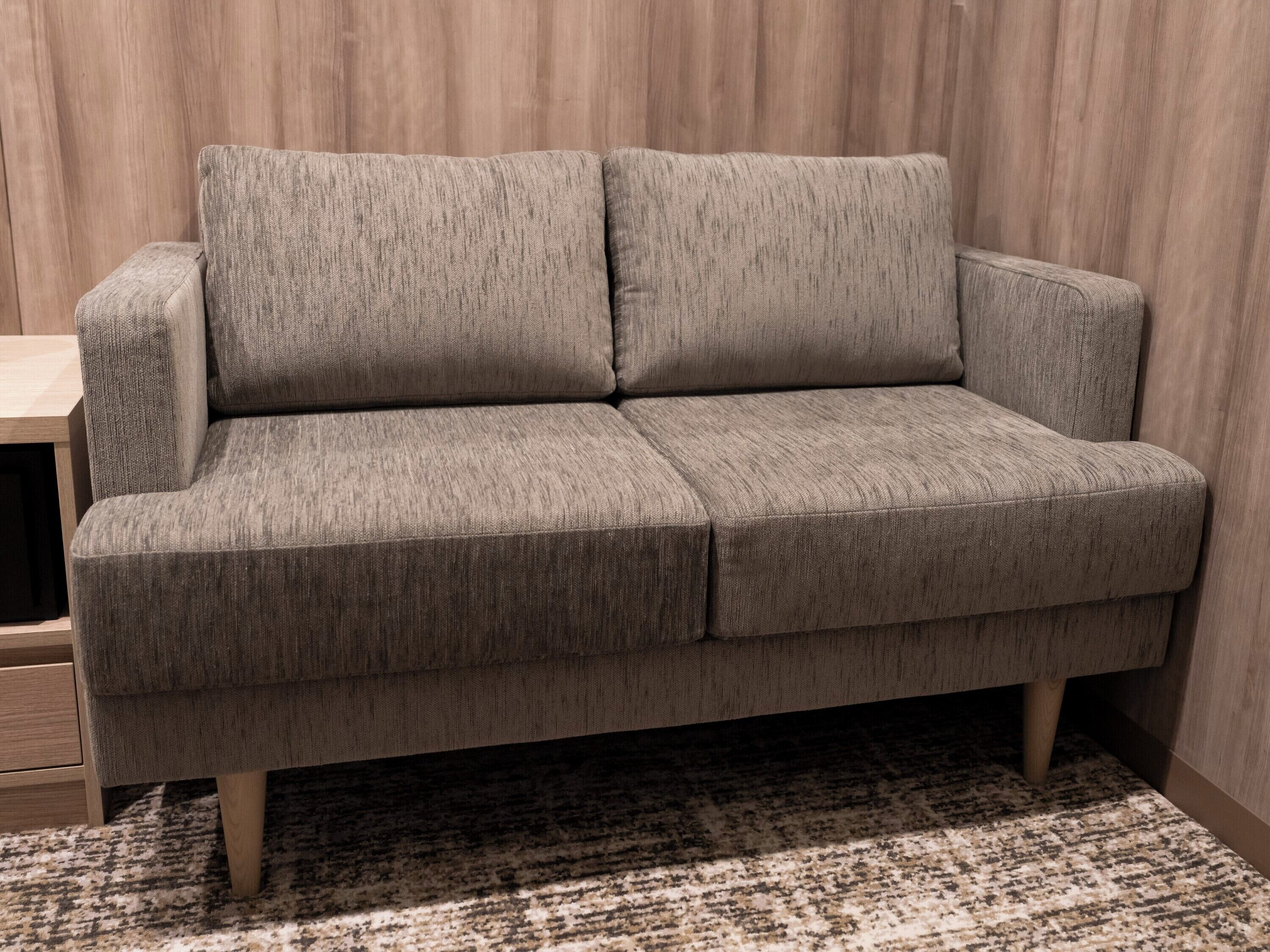A grey sofa with wooden legs, a patterned rug, and a wooden wall.