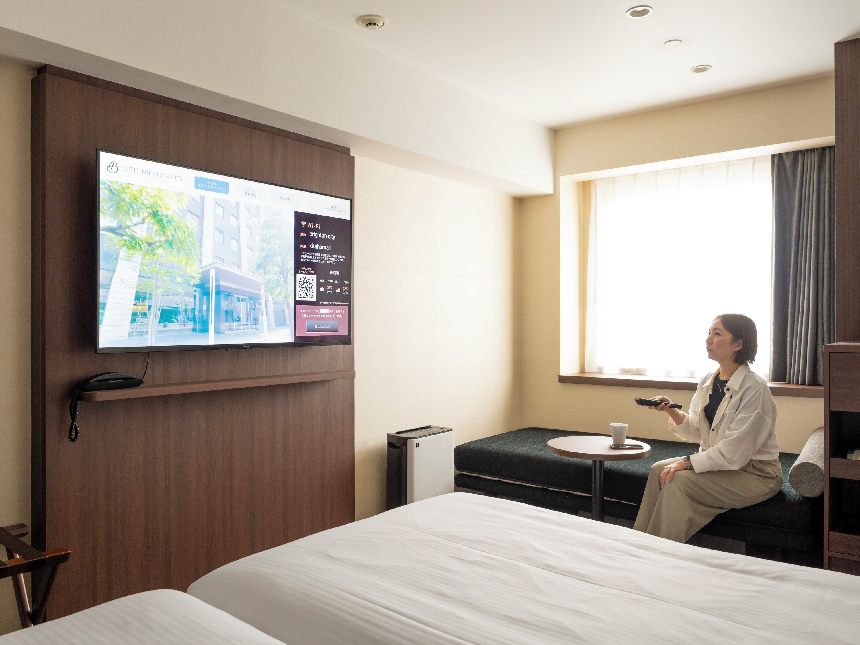 A hotel room with a bed, a desk, a chair, a TV mounted on the wall, and a window with curtains.
