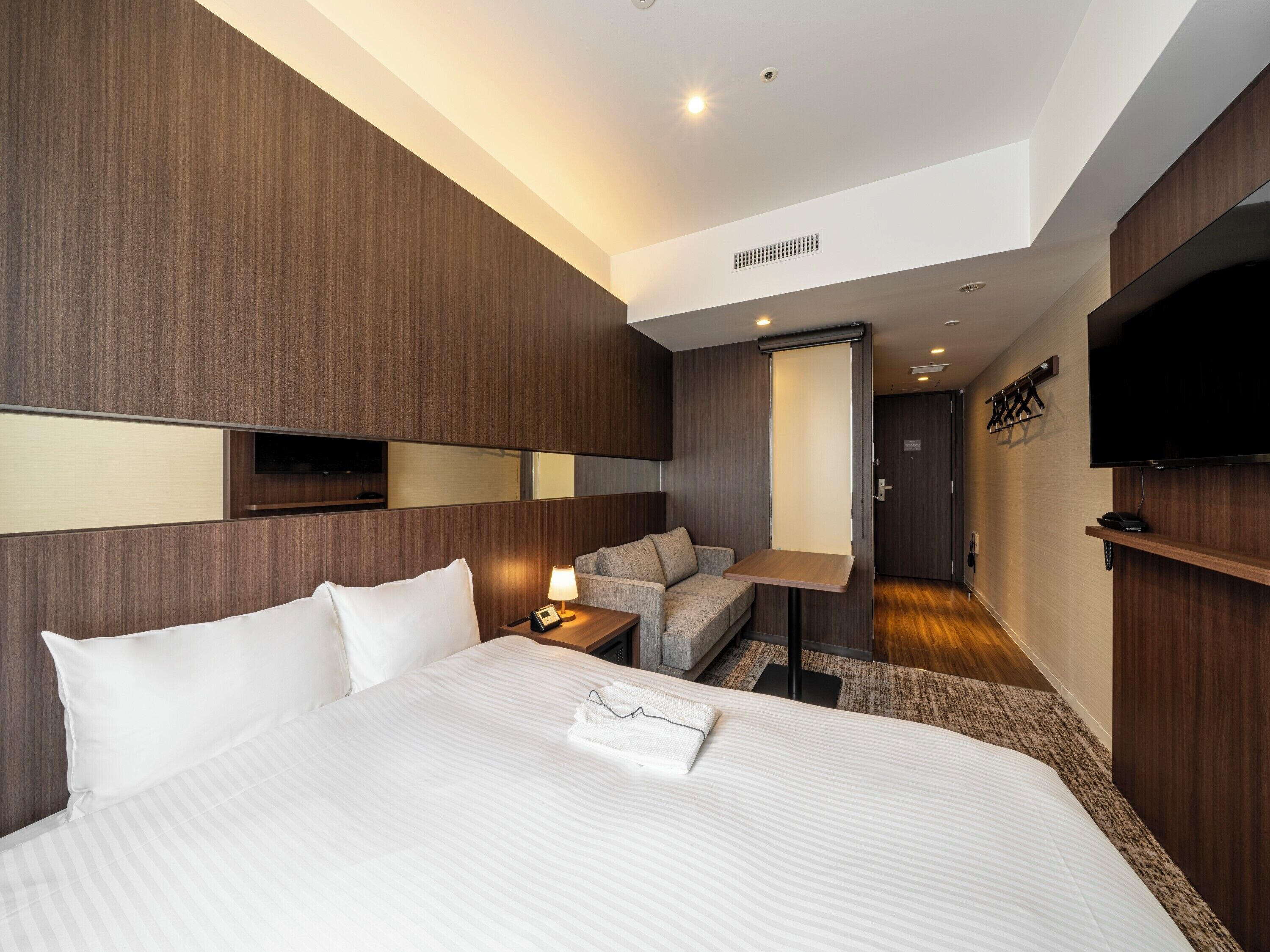 A modern hotel room with a large bed, a desk, a sofa, a TV, and a mirror.