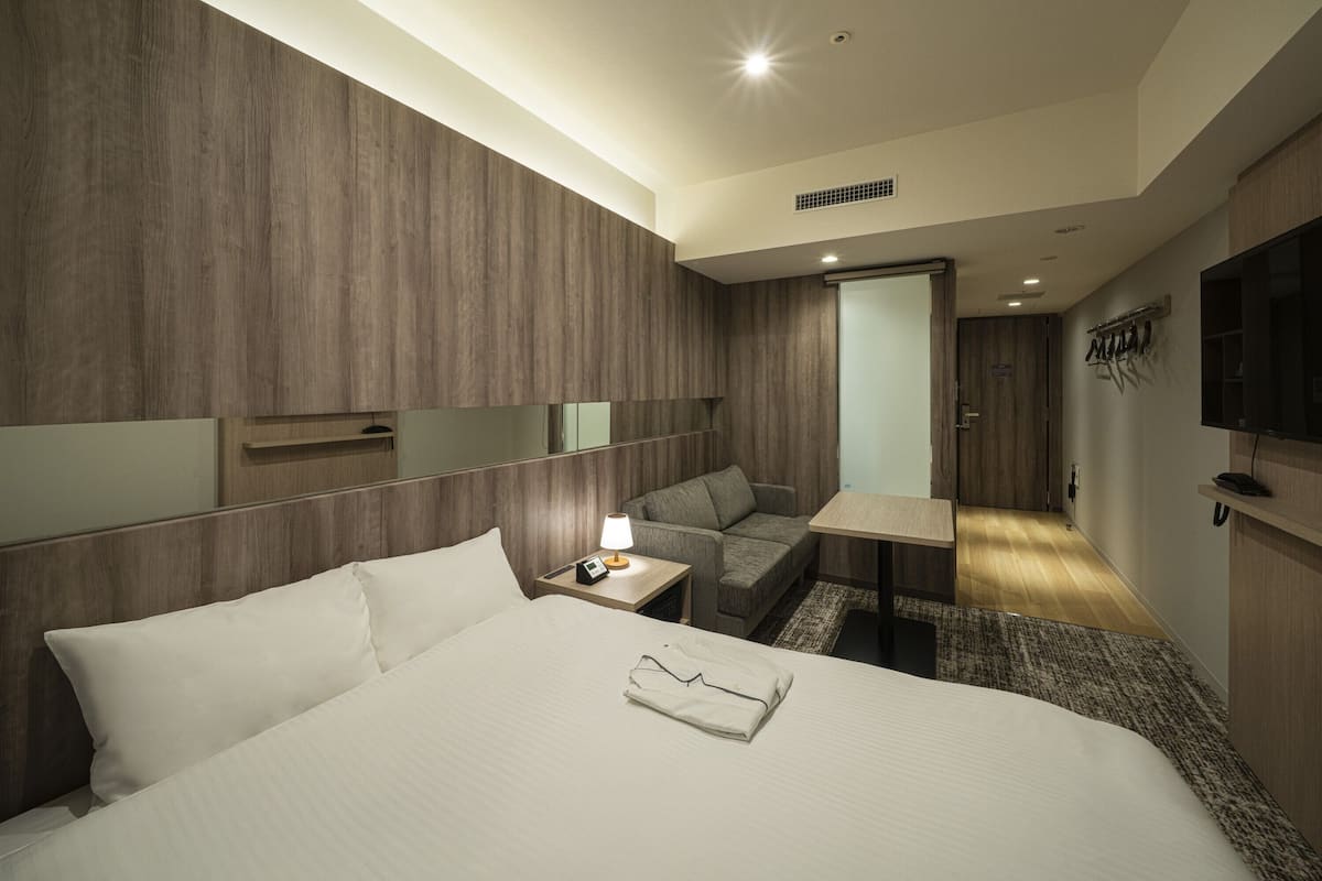 Een hotelkamer met een bed, nachtkastje, lamp en een zithoekje met een bank en een tafeltje.