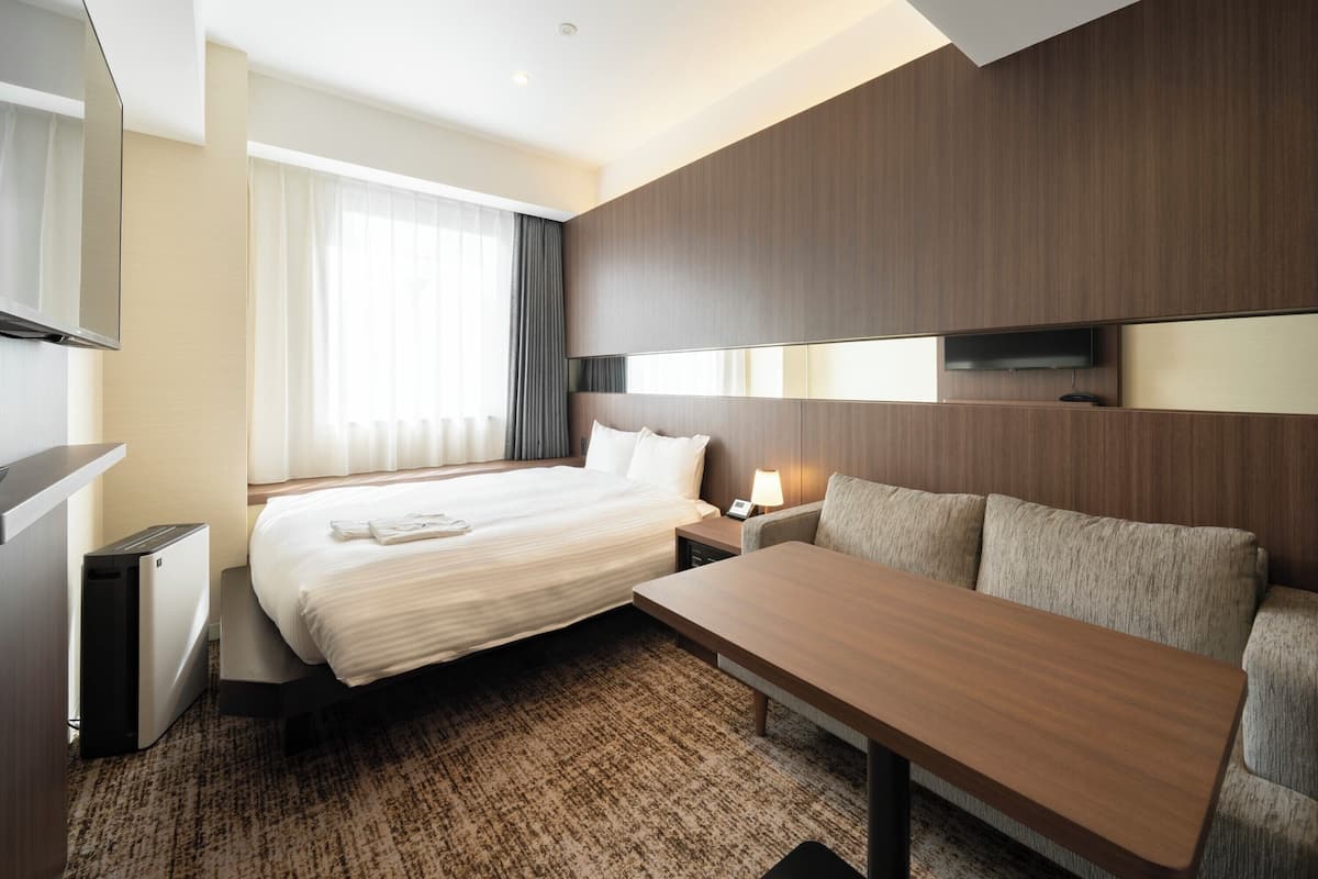 Hotelkamer met bed, nachtkastje, lamp, televisie en een zithoek met een bank en fauteuil.