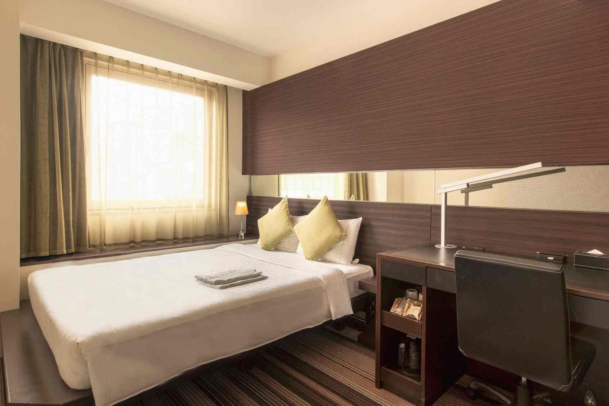 Een hotelkamer met een bed, nachtkastje, lamp en een groot raam met gordijnen.