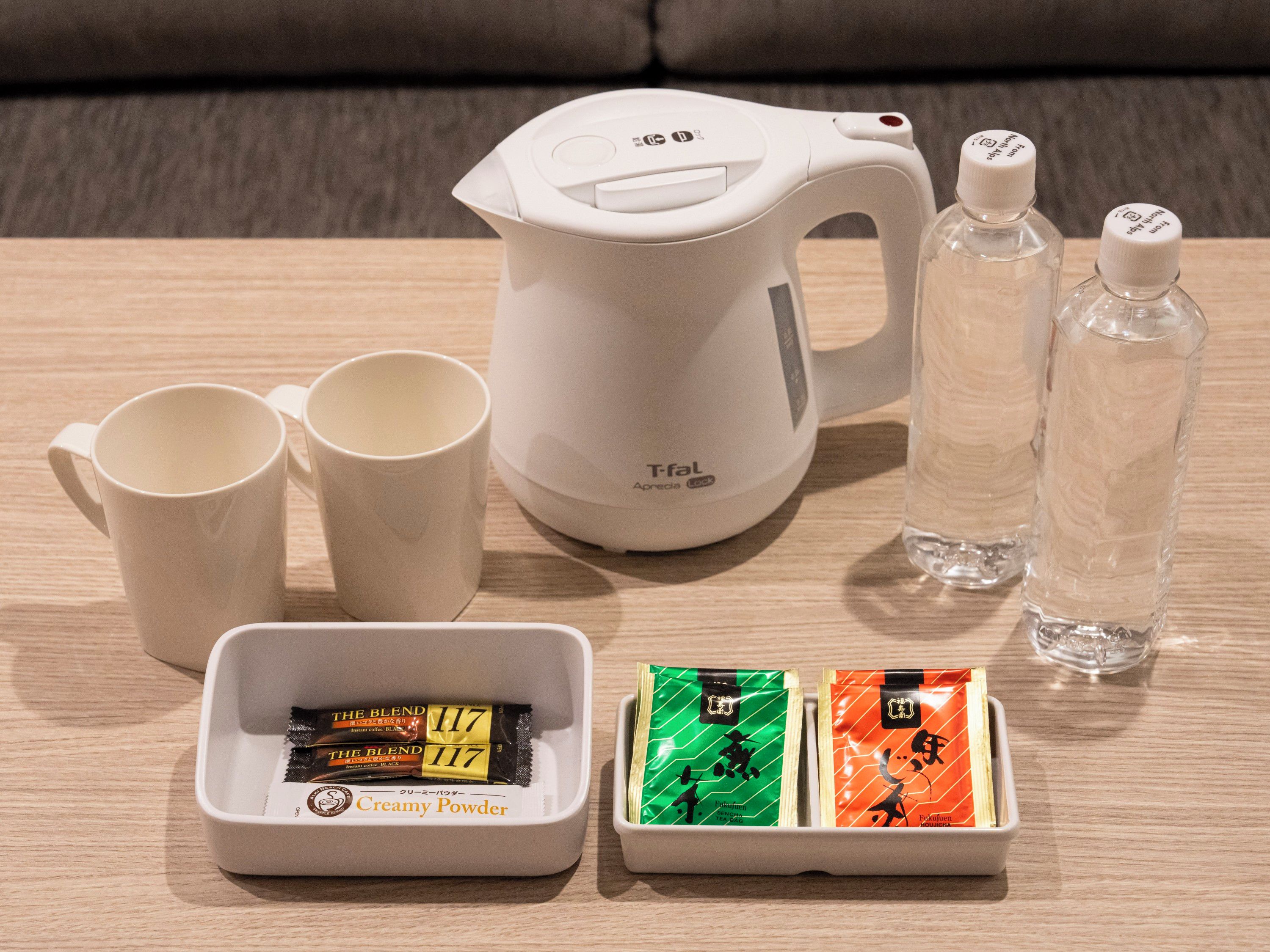 Una tetera eléctrica blanca, dos botellas de agua transparentes, dos tazas blancas y una bandeja con dos bolsitas de té y una bolsita de crema en polvo.