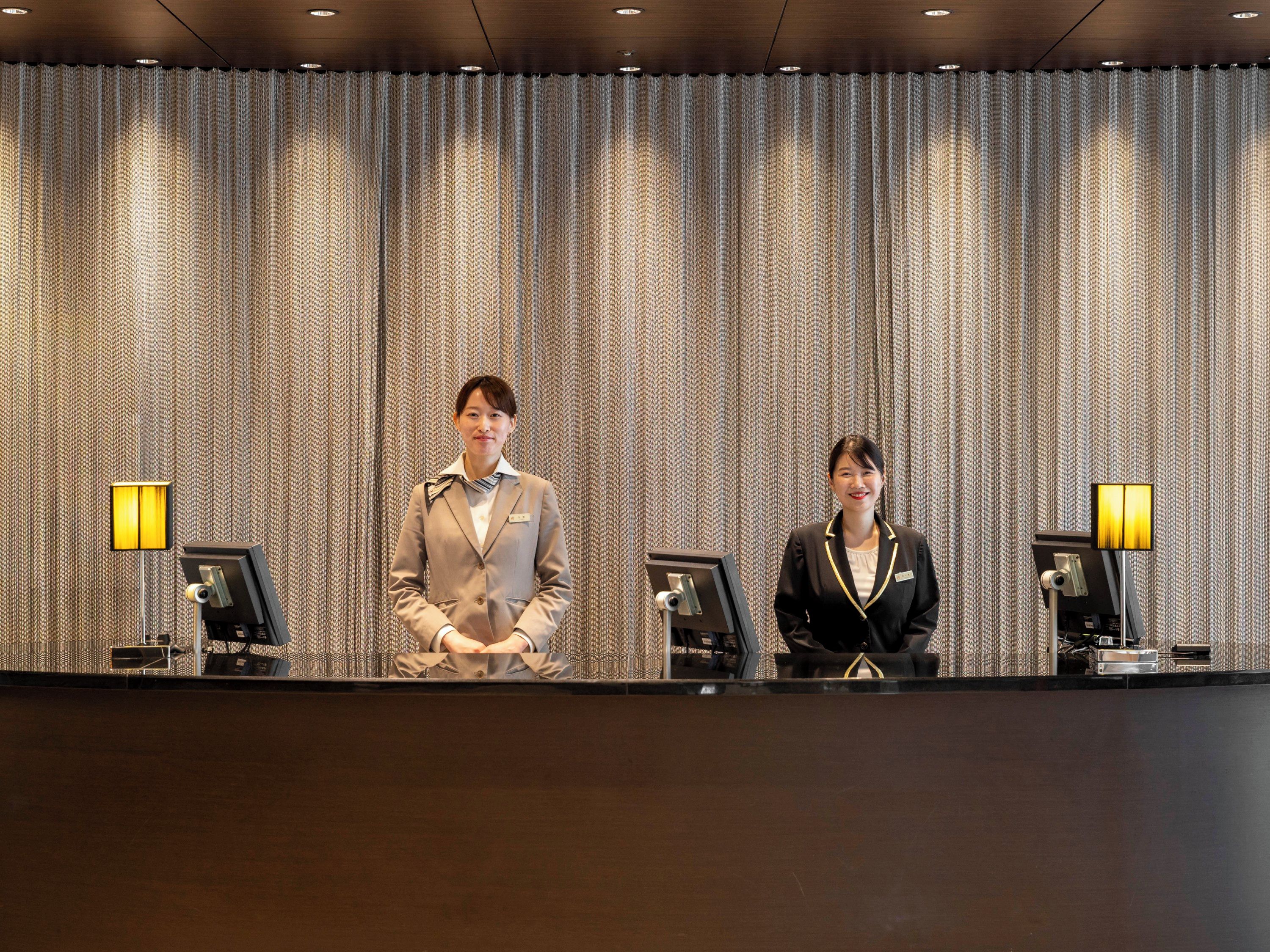 Two receptionists standing behind a reception desk with a striped curtain backdrop and desk lamps.