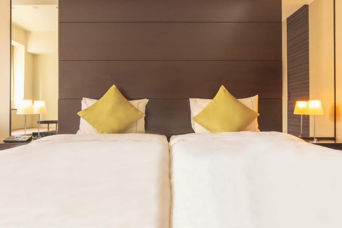 Een tweepersoonsbed met witte lakens en twee gele kussens, een donker houten hoofdbord en twee bedlampen met witte lampenkappen.