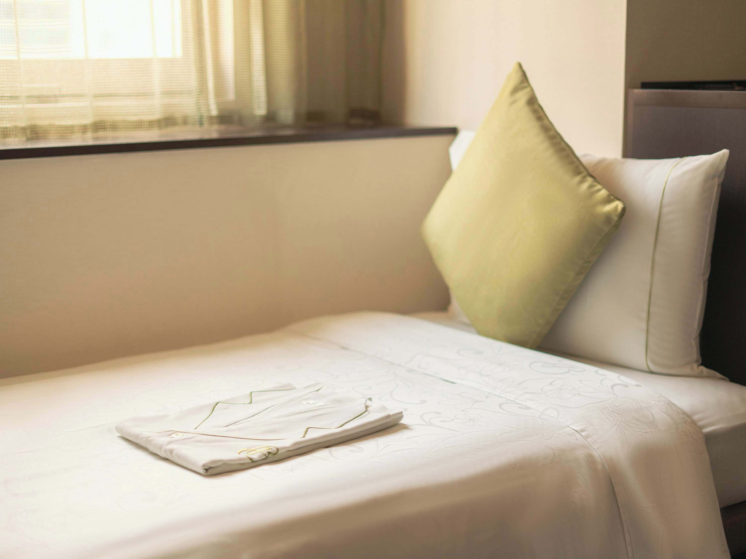 Ein ordentlich bezogenes Bett mit einem gefalteten Kleidungsstück, einem grünen Kissen und einem Fenster mit transparenten Vorhängen.