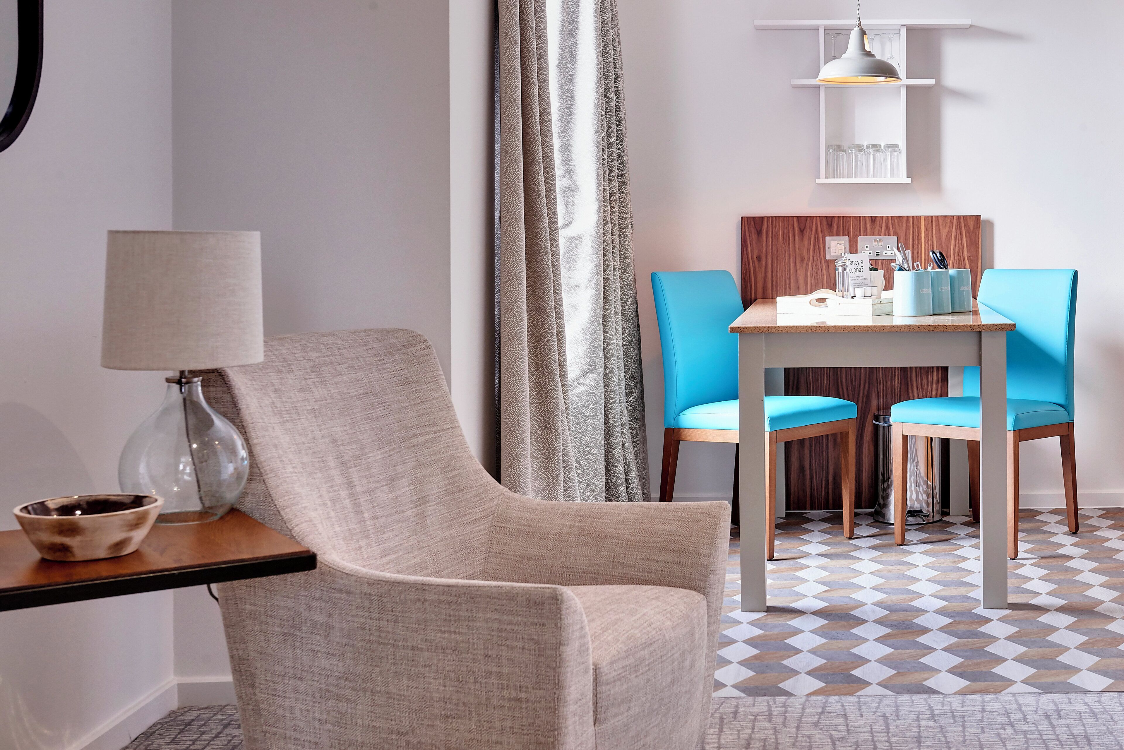 A modern room with a beige armchair, a small table with a lamp, a blue chair, and a wooden dining table with chairs.