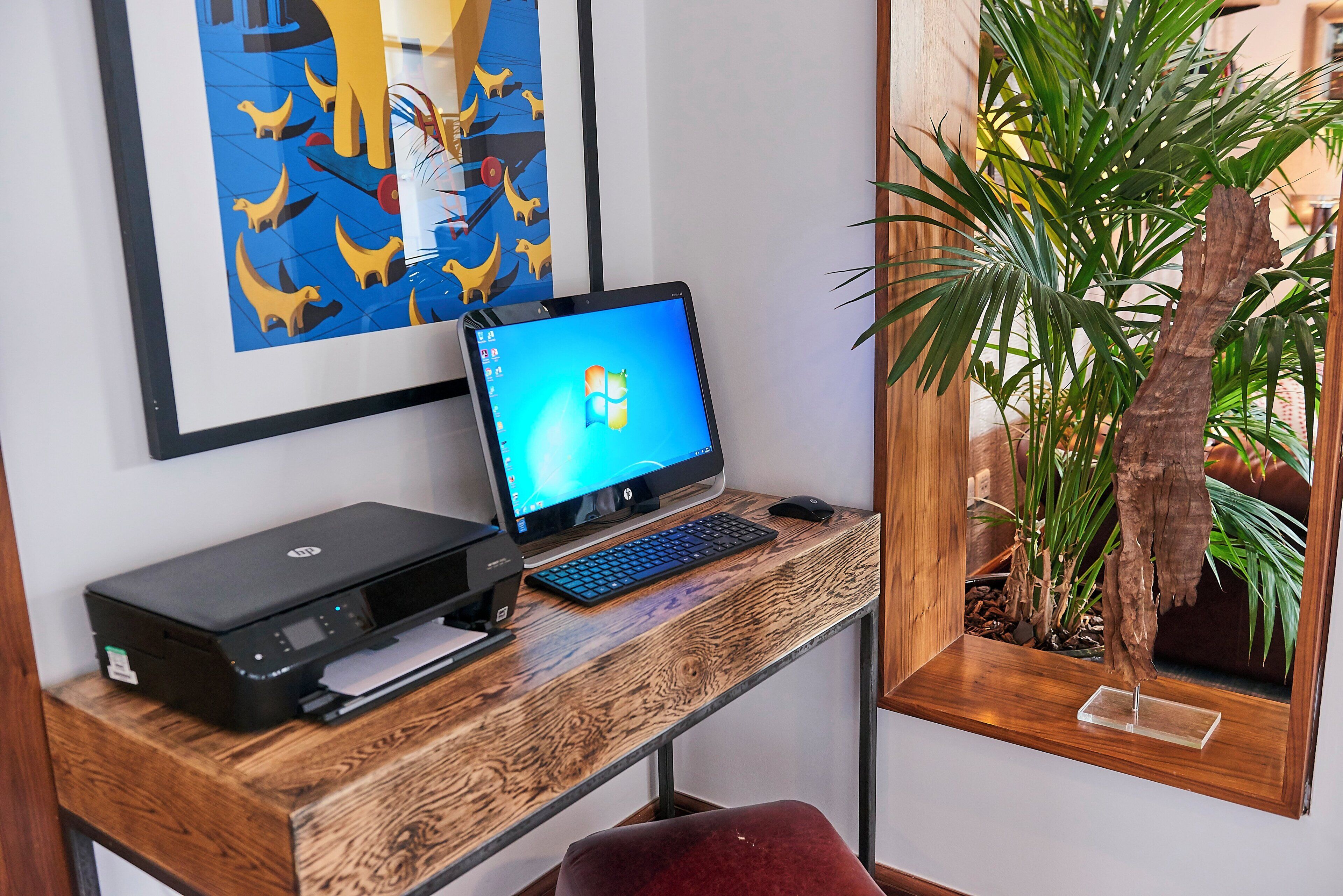 A wooden desk with an HP desktop computer, a keyboard, and a printer. There is a framed artwork on the wall and a potted plant on the desk.