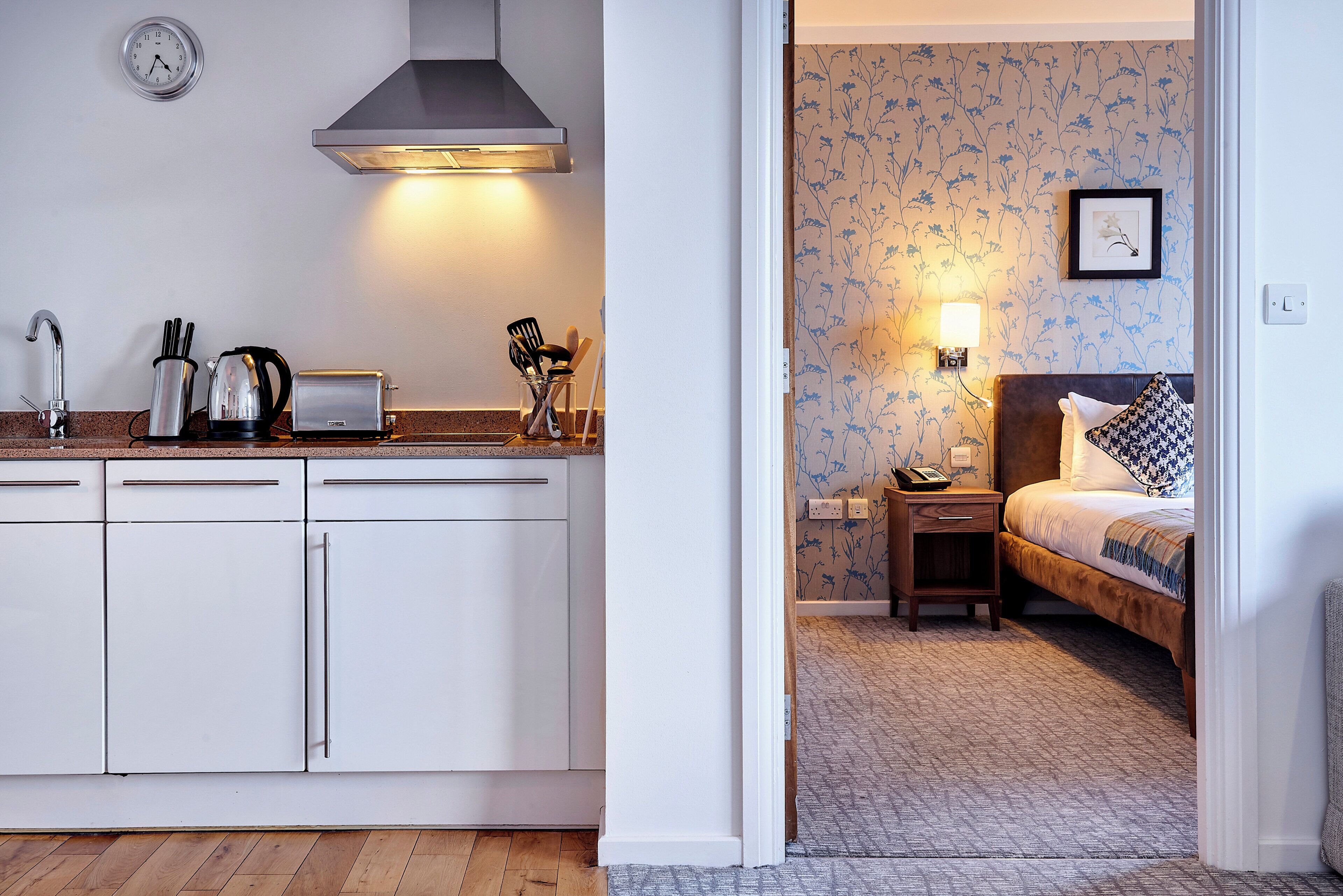 Een moderne keuken met witte kasten, een broodrooster en een waterkoker. Er hangt een klok aan de muur en een lamp boven het fornuis.