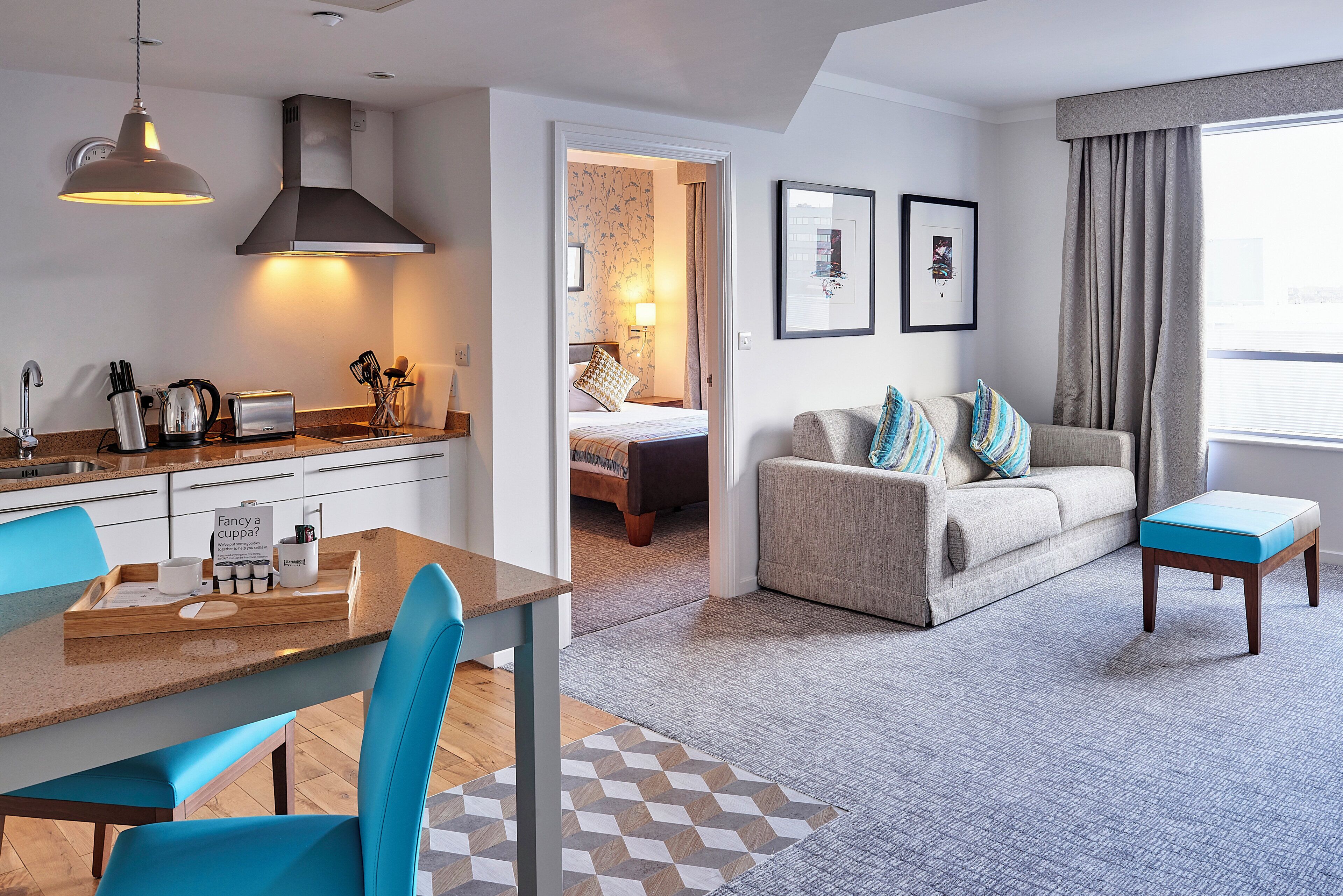A modern hotel room with a kitchenette, a dining area, a sofa with cushions, and a large window with curtains.