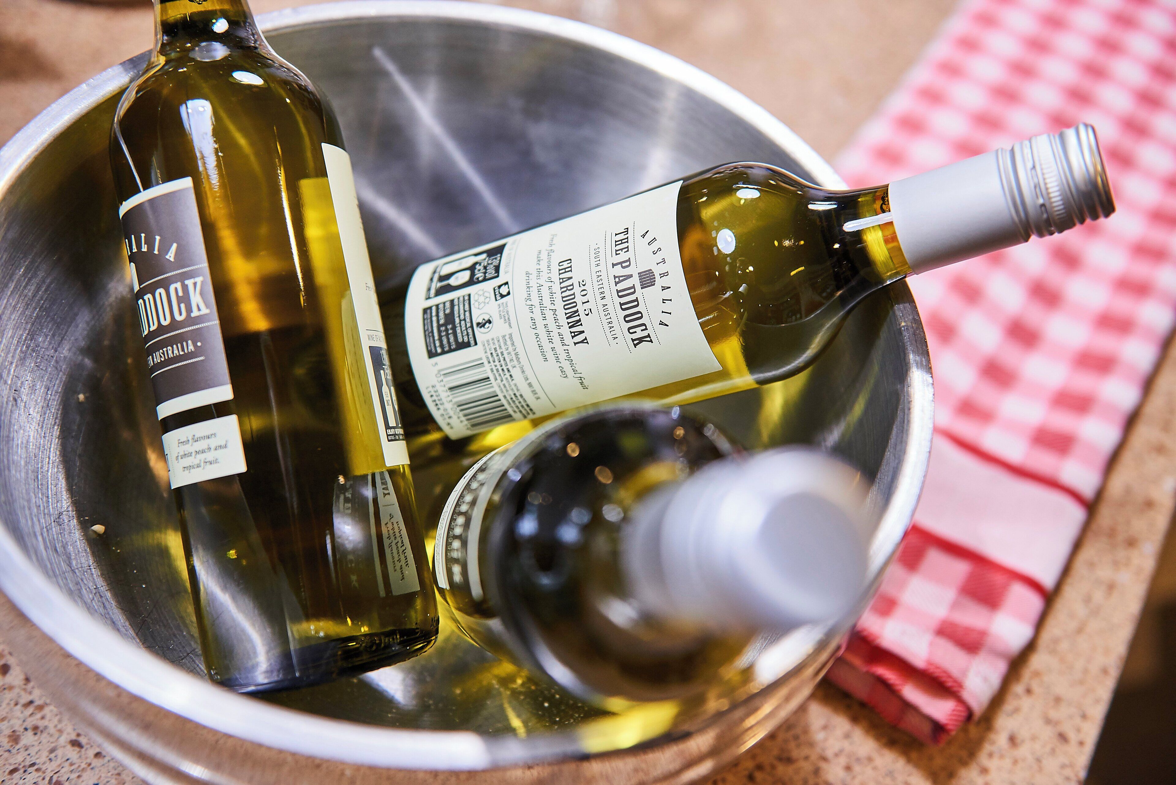 Two wine bottles in a metal container with a wine opener and a checkered cloth.
