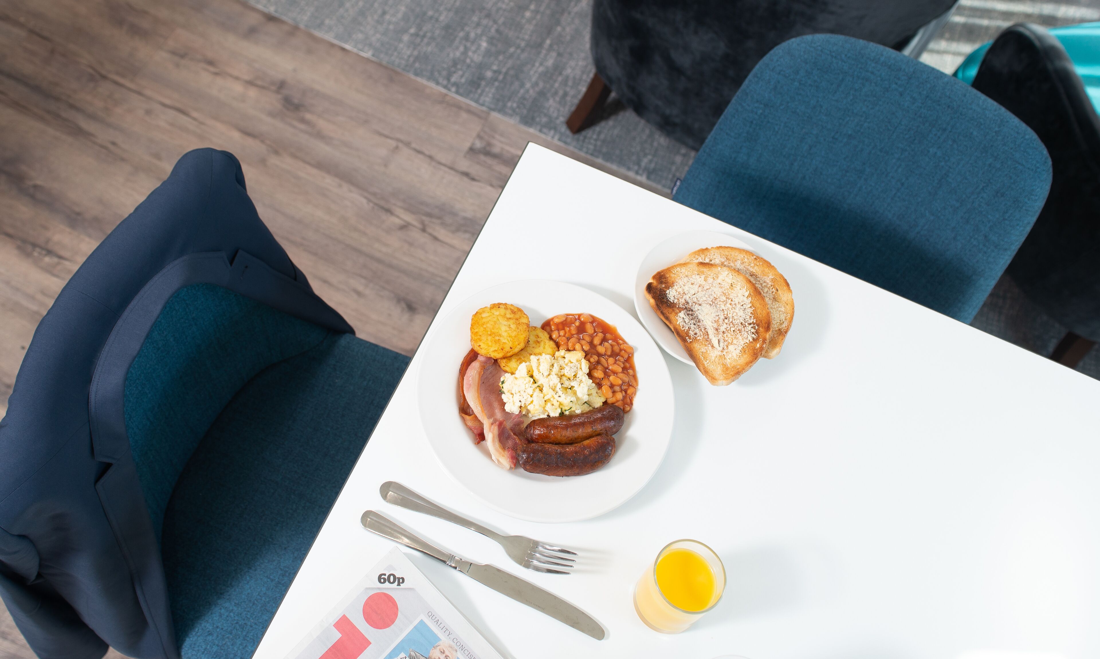 A meal with toast, beans, sausages, and scrambled eggs on a white plate, accompanied by a glass of juice and cutlery on a table.